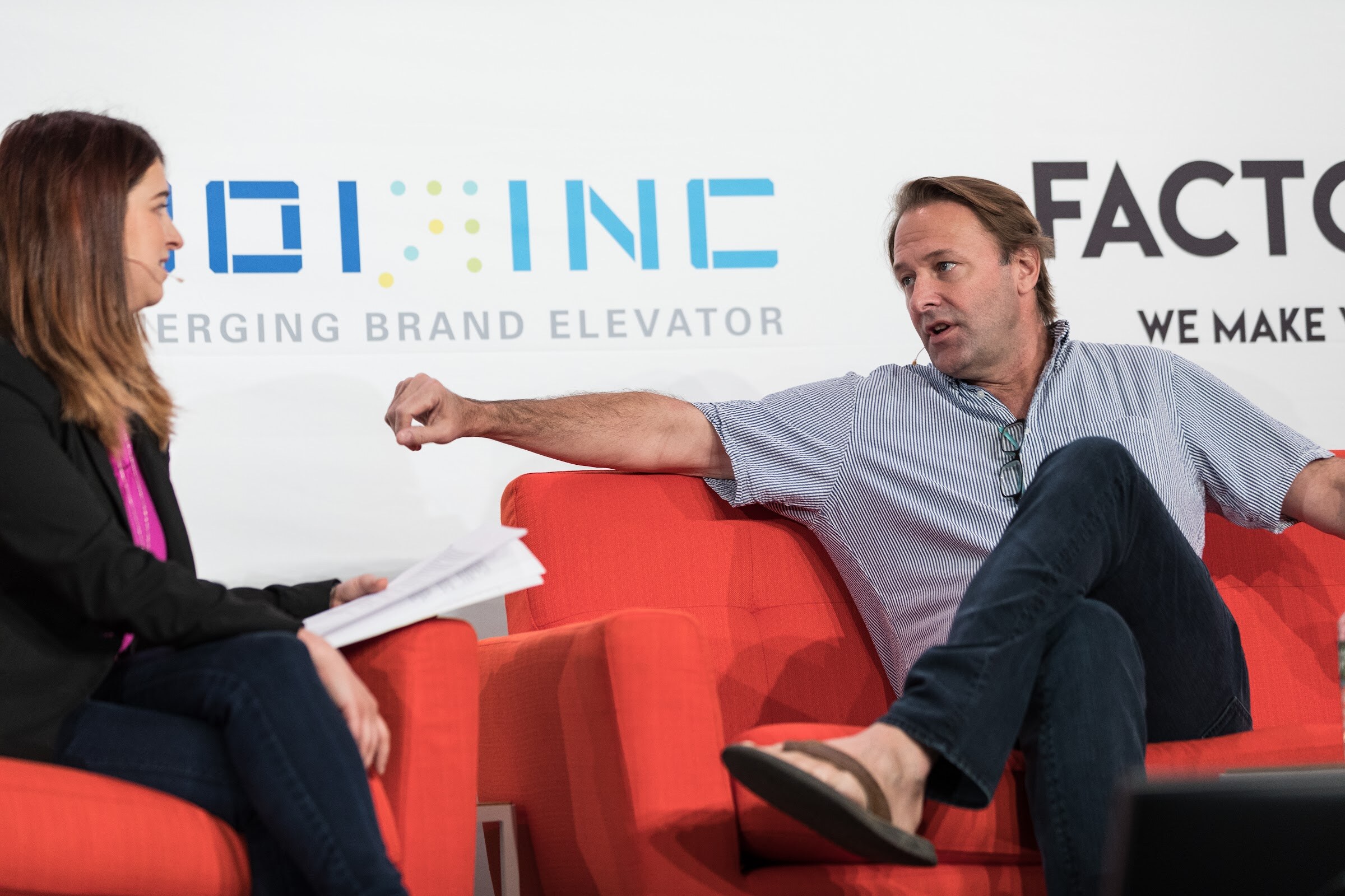 A man in a striped short-sleeve shirt and sandals sits sideways on an orange sofa, arm extended, talking to a woman with notes in the foreground.
