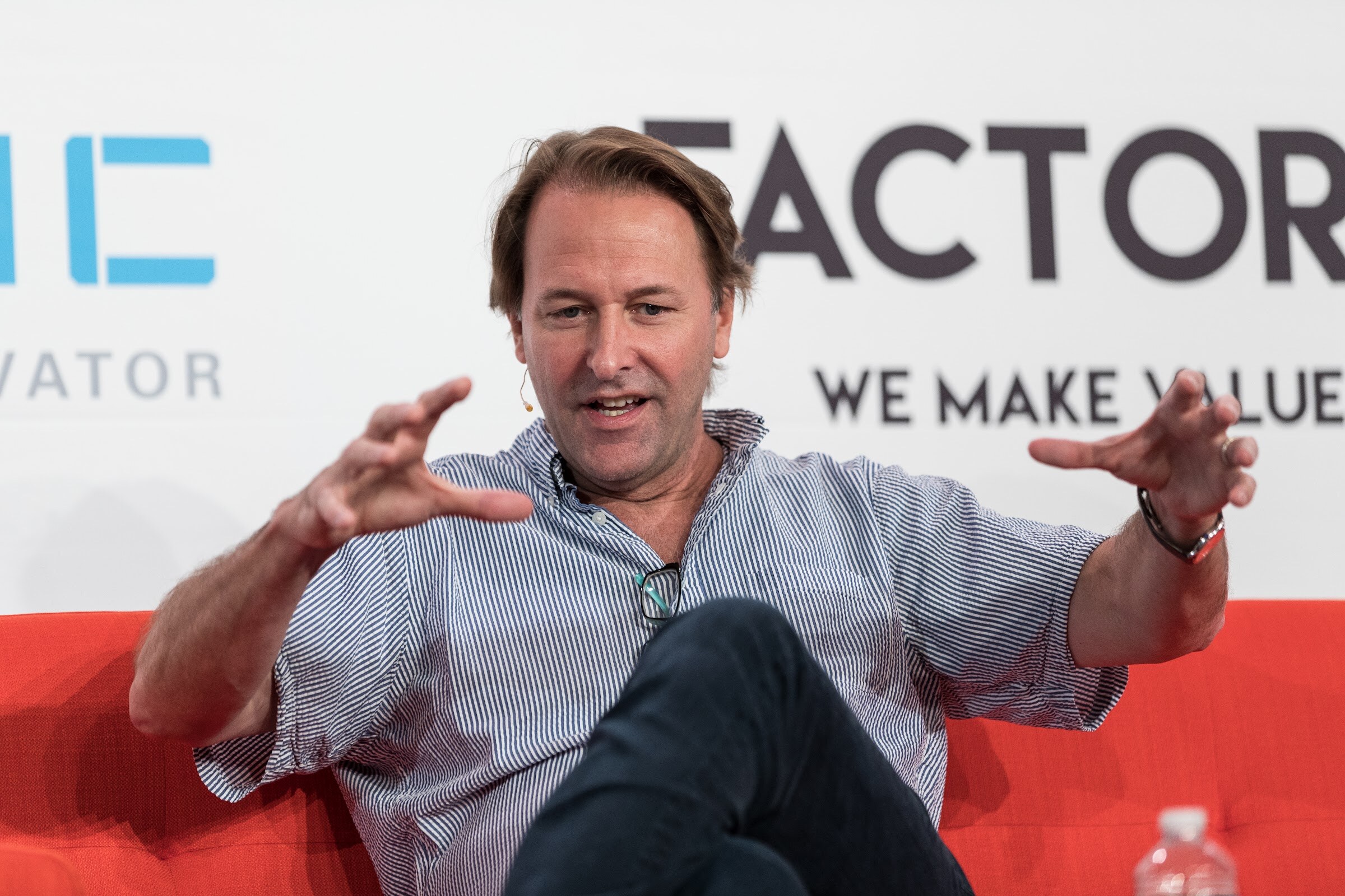 A man in a blue striped shirt sits on an orange sofa with both hands stretched wide as he speaks, a water bottle at the lower right.