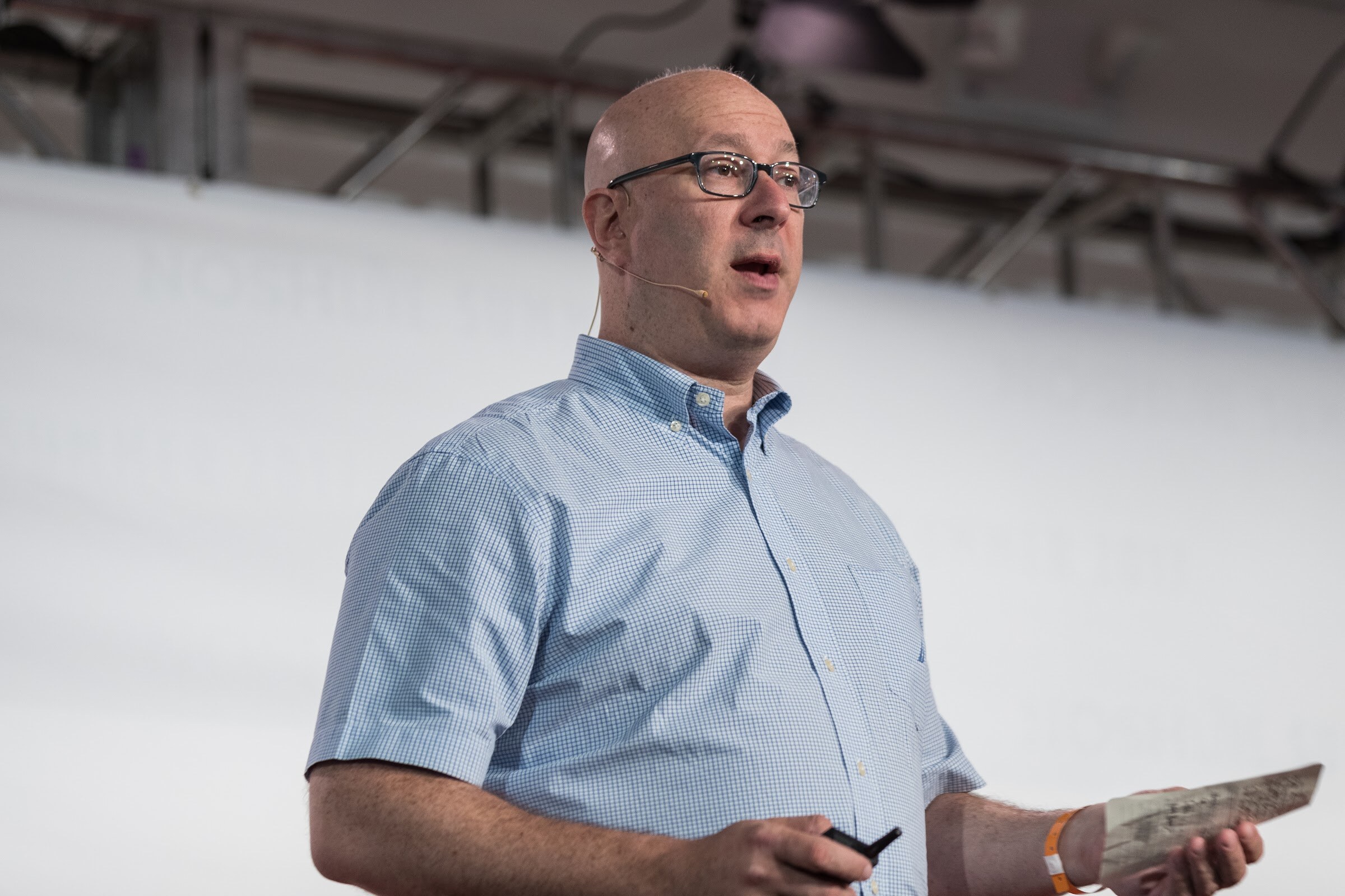 A bald man with glasses and a headset microphone, in a light blue short-sleeve shirt, holds a clicker and a folded paper against a pale stage backdrop.