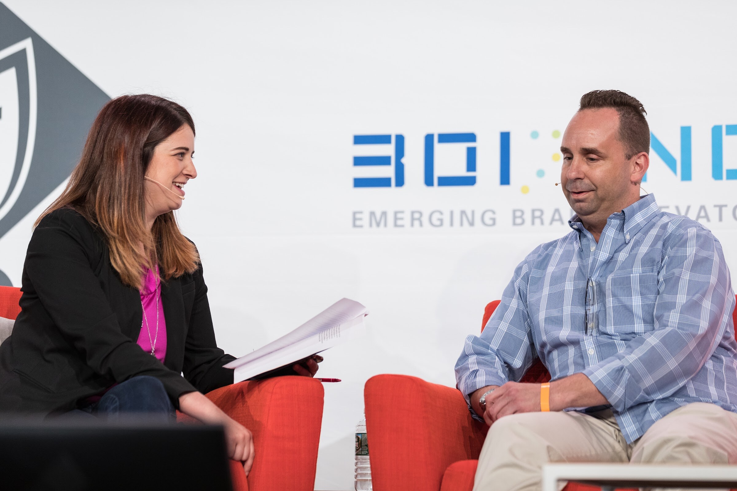 A woman in a black blazer and magenta top smiles and holds papers across from a smiling man in a blue plaid shirt on orange armchairs.
