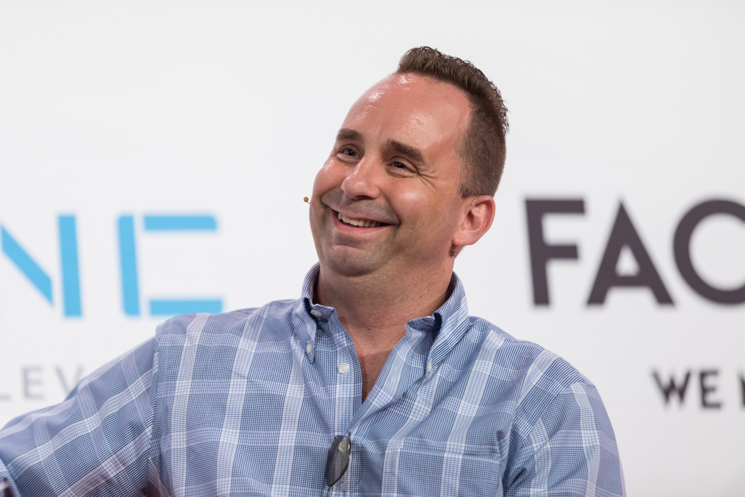 A close view of a smiling man in a blue plaid button-down shirt wearing a headset microphone, with a 301 Inc and Factory banner blurred behind him.