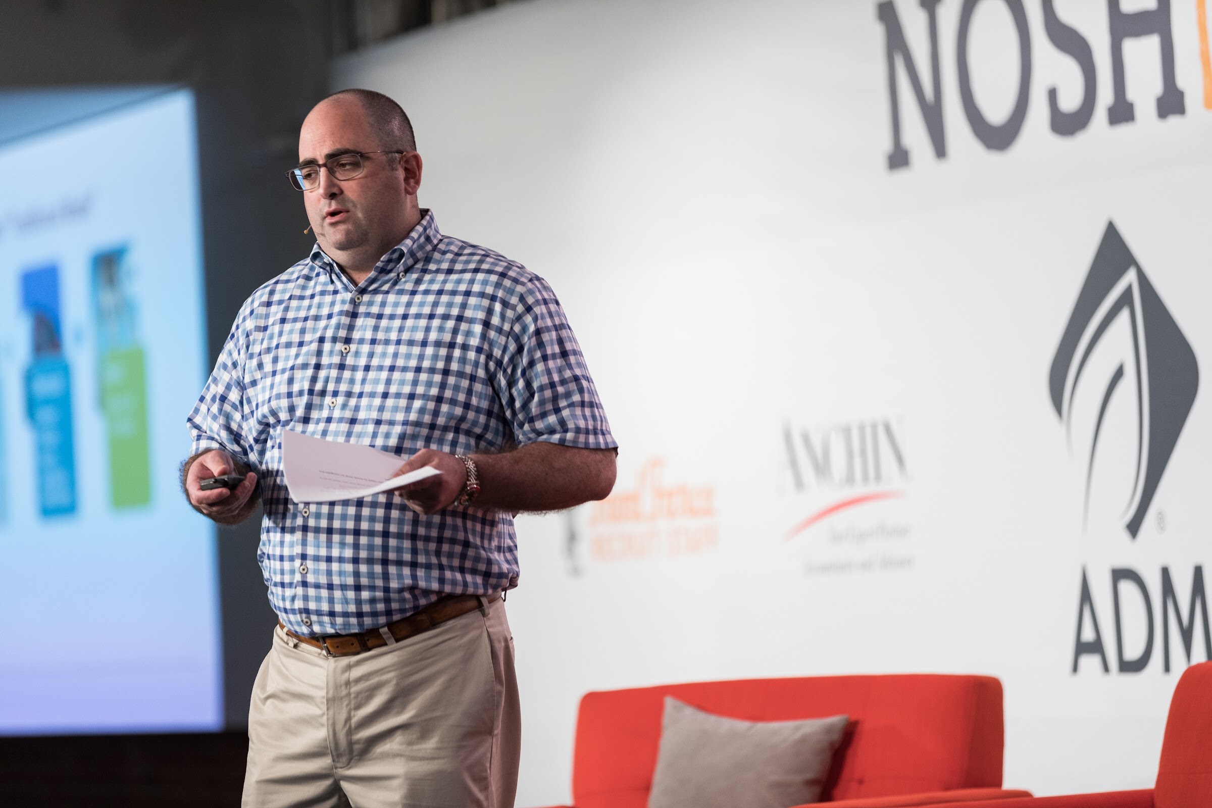 A man in glasses, a blue checked shirt and khakis holds a paper and a clicker on stage in front of ADM and Nosh Live signage, with a slide at left.