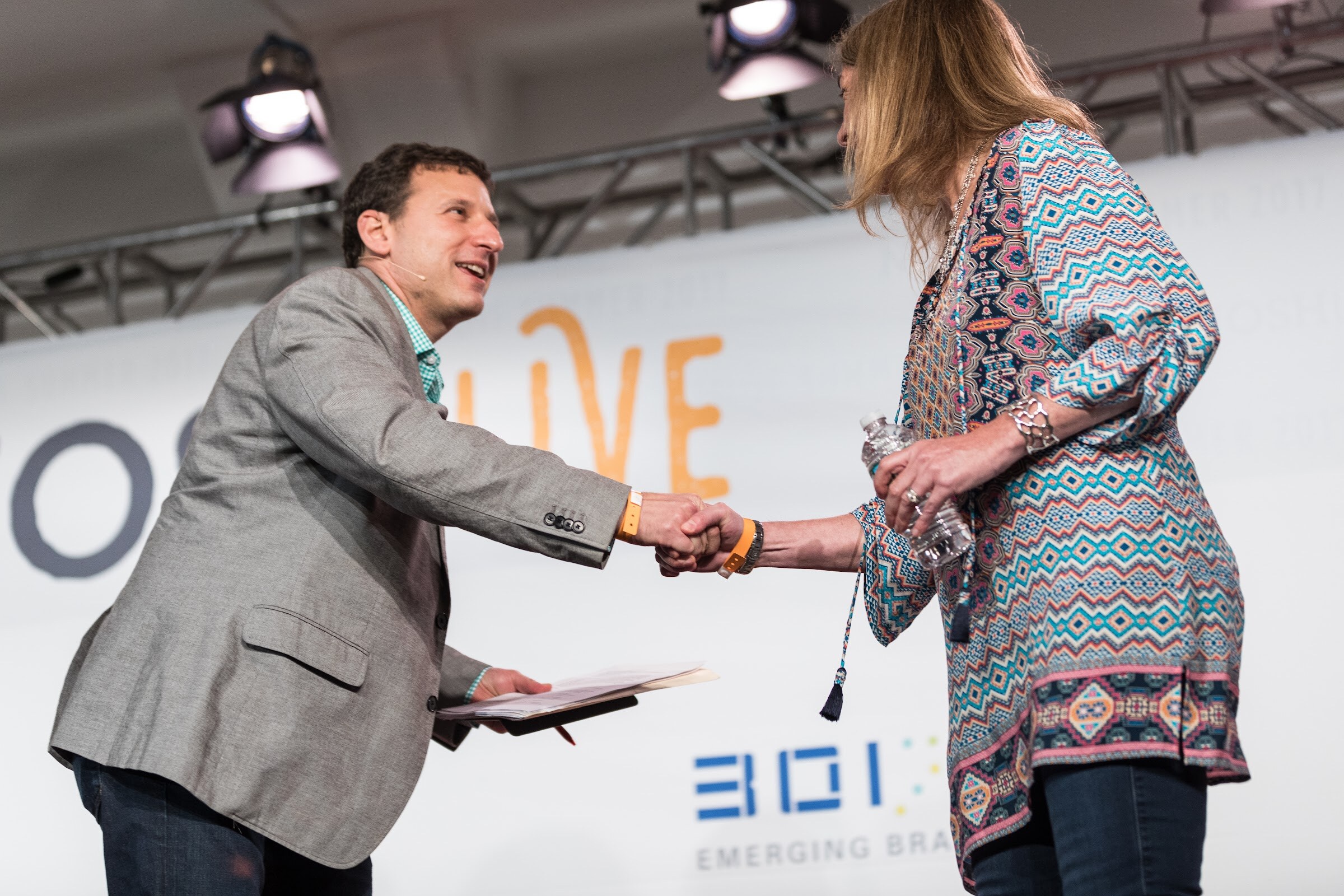 A man in a gray blazer holding papers shakes hands with a woman in a patterned tunic who holds a water bottle, under stage lighting.