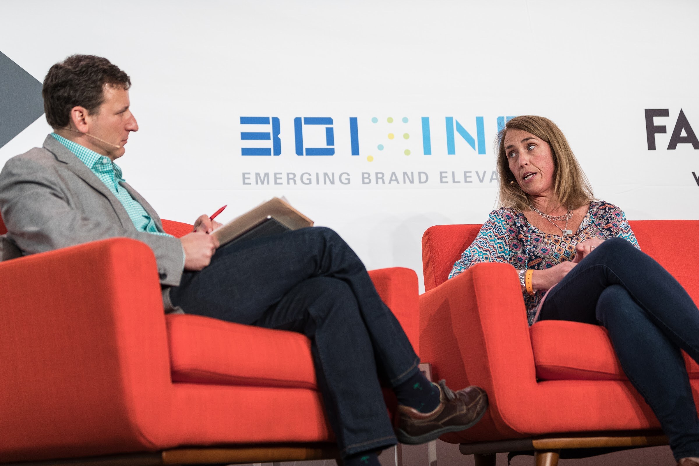 A woman in a patterned top sits in an orange armchair, looking toward a man in a gray blazer with notes and a red pen, with a 301 Inc banner behind.