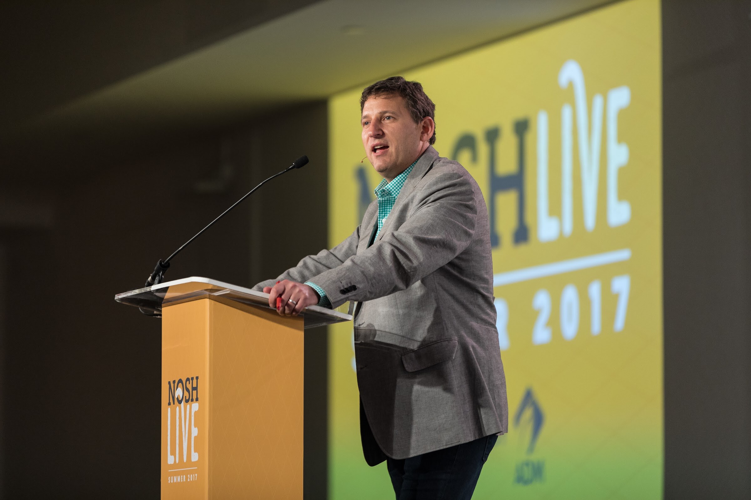 A man in a gray blazer leans on an orange Nosh Live podium in front of a yellow projection screen reading Nosh Live Summer 2017.