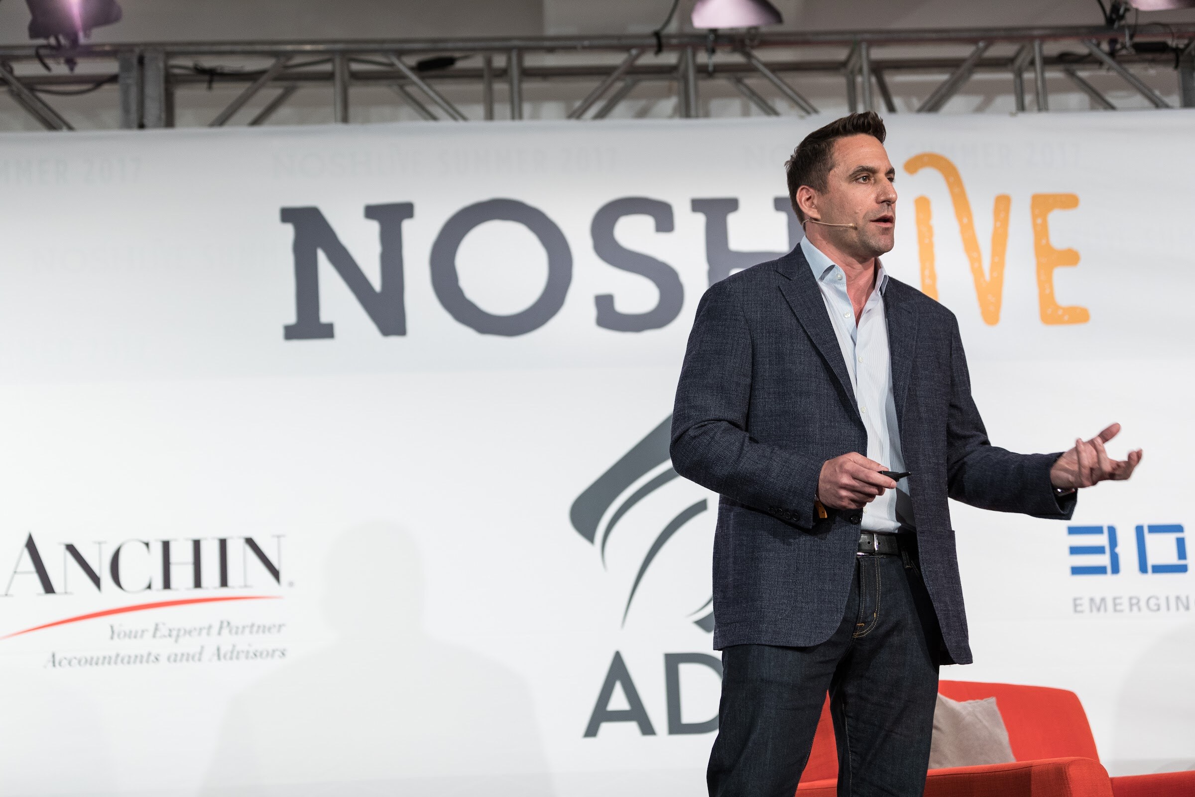 A man in a navy blazer and open collar stands on stage with one hand extended in front of a Nosh Live backdrop with Anchin and ADM logos.