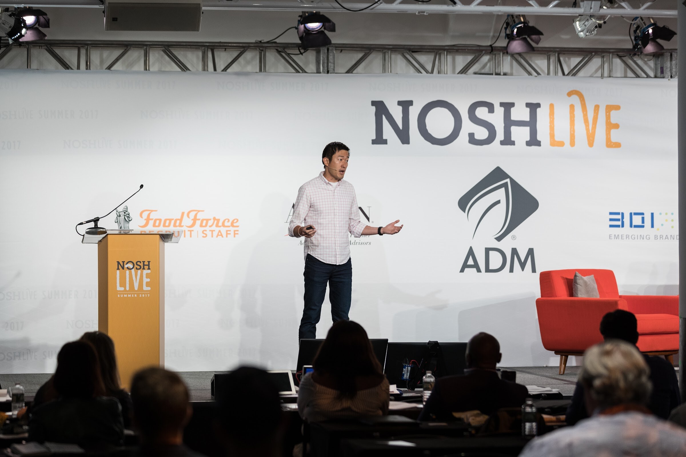 A man in a pink checked shirt and dark jeans stands on a stage in front of a large Nosh Live and ADM backdrop, with an orange podium at left and audience heads below.