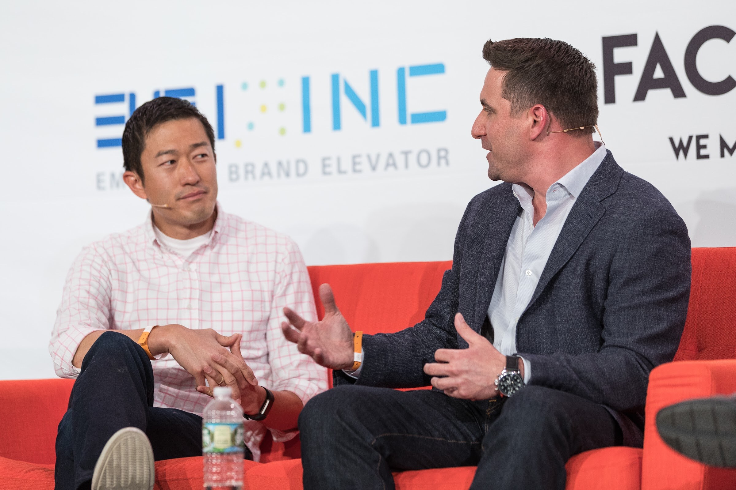 Two men sit on an orange sofa, one in a pink checked shirt and one in a navy blazer gesturing, in front of a Factory banner.