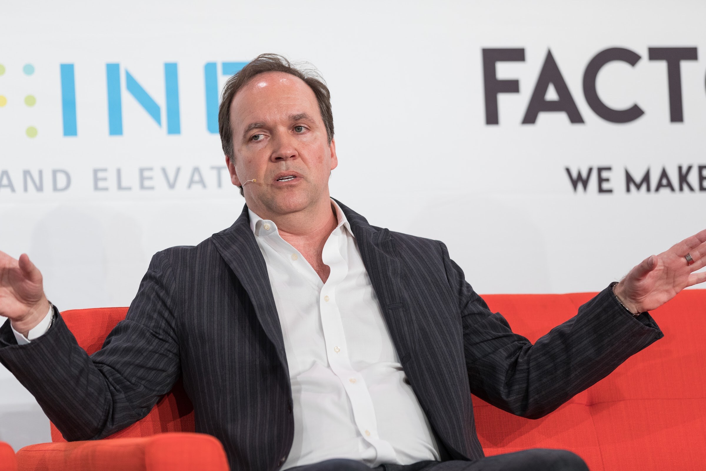 A man in a dark pinstriped jacket and white open-collar shirt sits on an orange sofa with both arms spread to the sides.