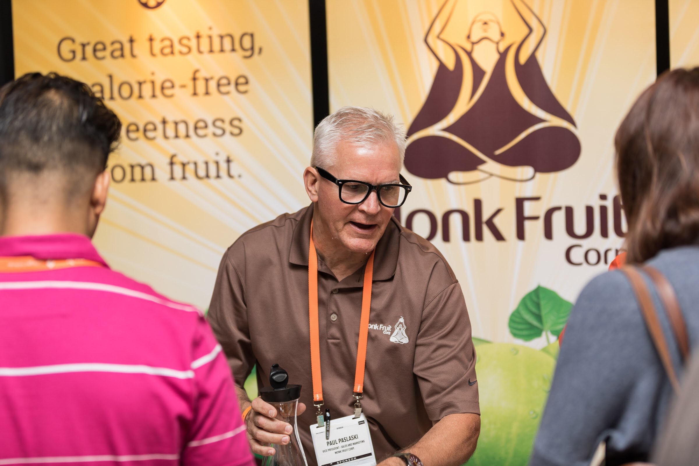 A man with glasses in a brown polo and orange lanyard speaks to visitors in front of a Monk Fruit Corp. banner.