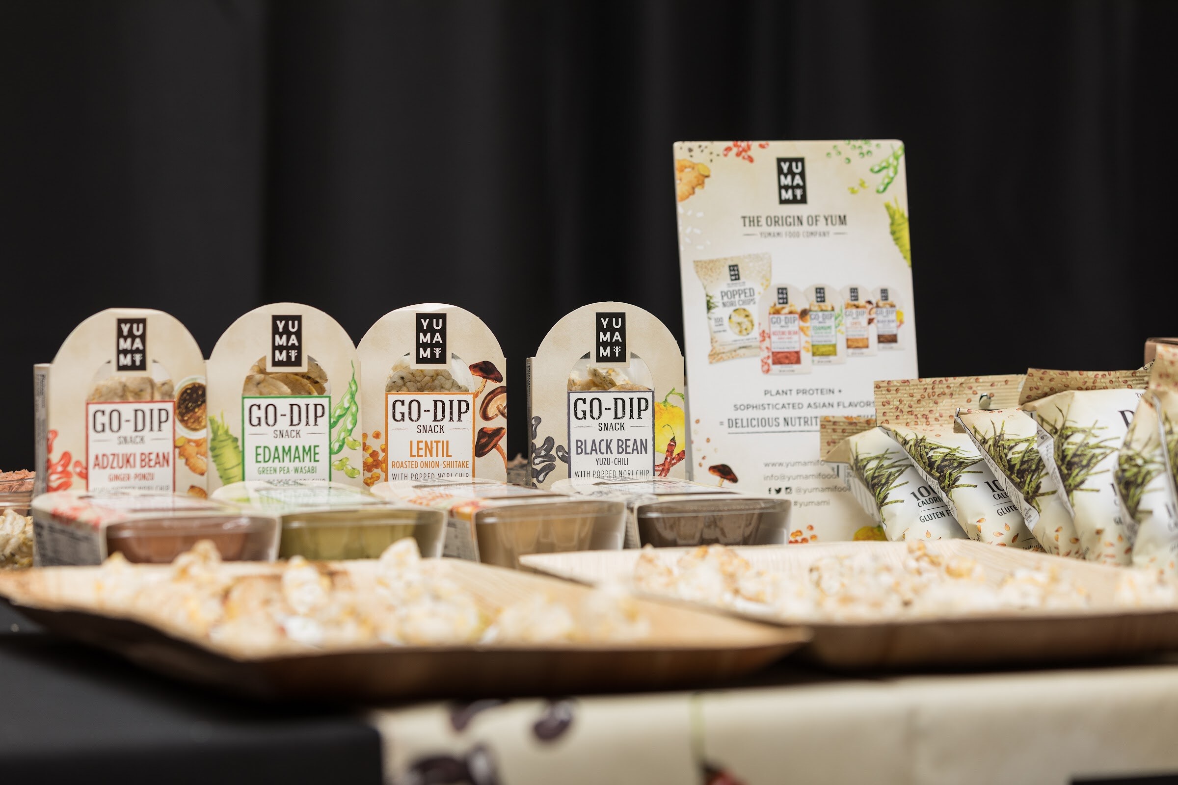 A table display of Go-Dip snack packs in Adzuki Bean, Edamame, Lentil and Black Bean flavors, with sampling trays and a Yumami sign.