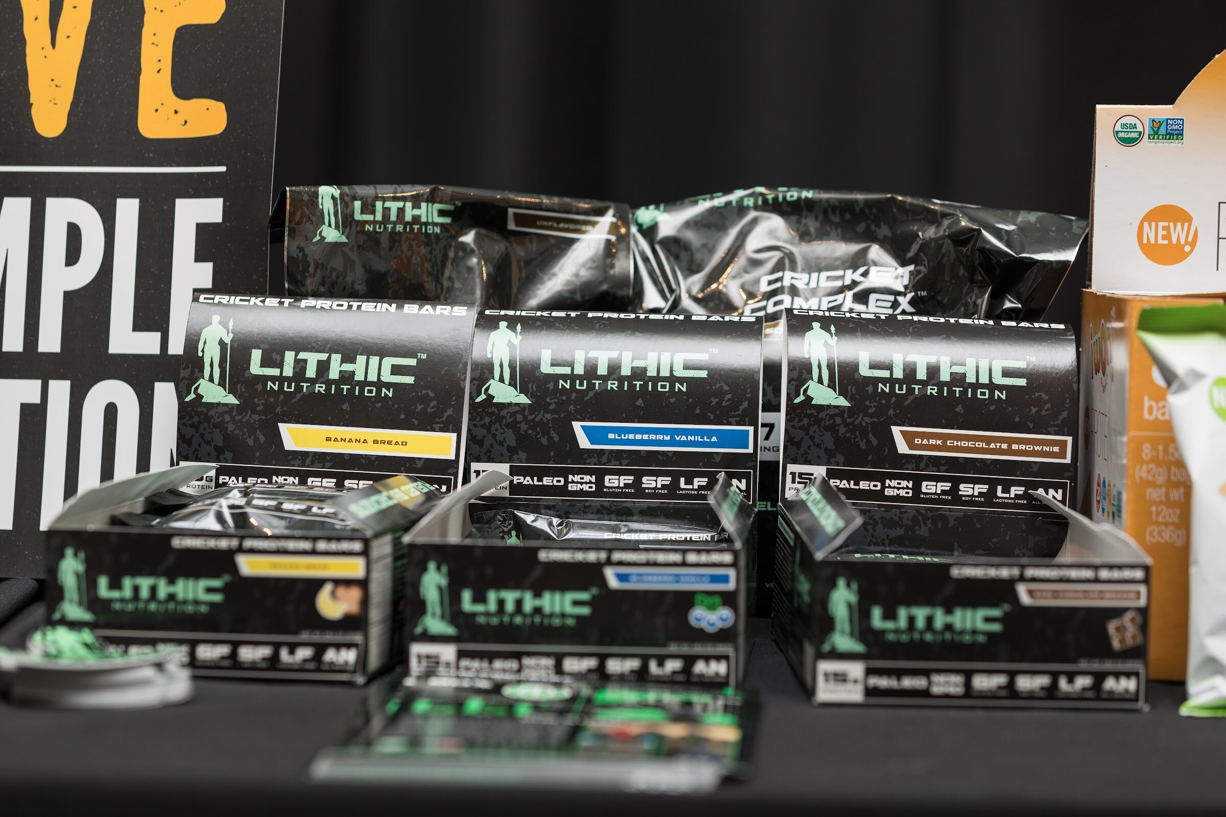 Black boxes of Lithic Nutrition cricket protein bars in banana bread, blueberry vanilla and dark chocolate brownie flavors on a black table.