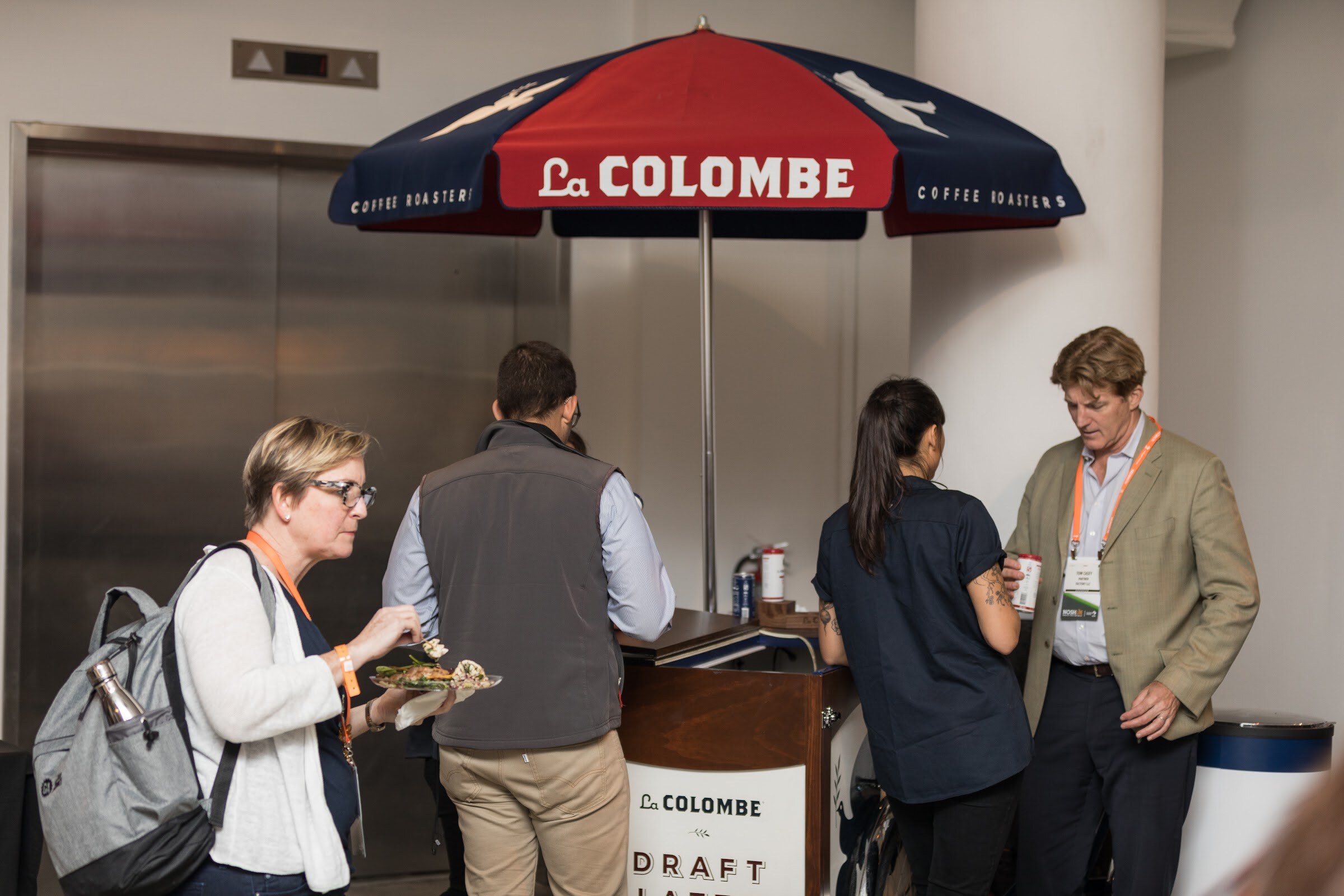 A cart under a red and navy umbrella reading La Colombe Coffee Roasters, with attendees holding plates and cups beside it.