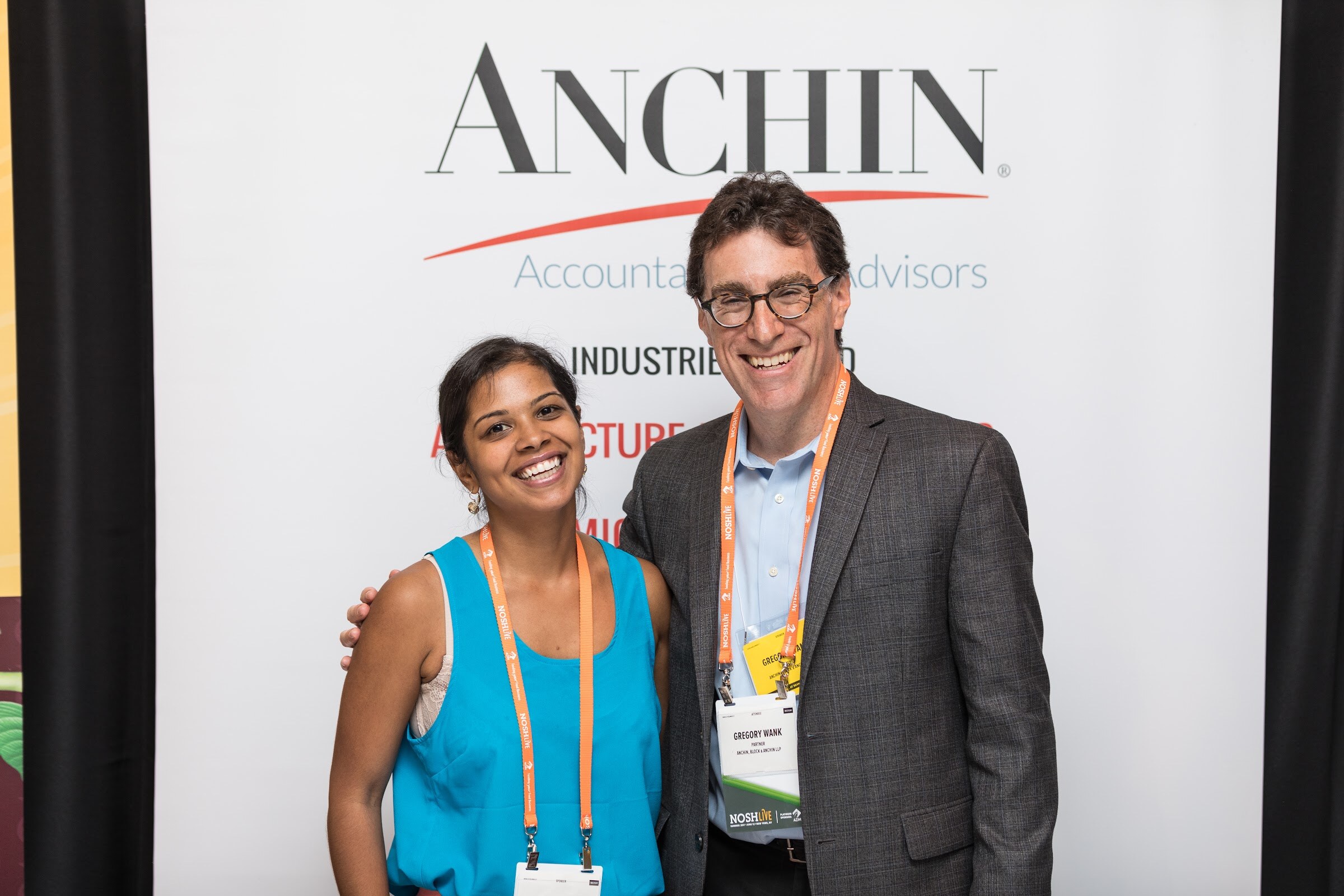 A smiling woman in a turquoise top and a man in a gray blazer, both wearing orange lanyards, pose in front of a white Anchin banner.