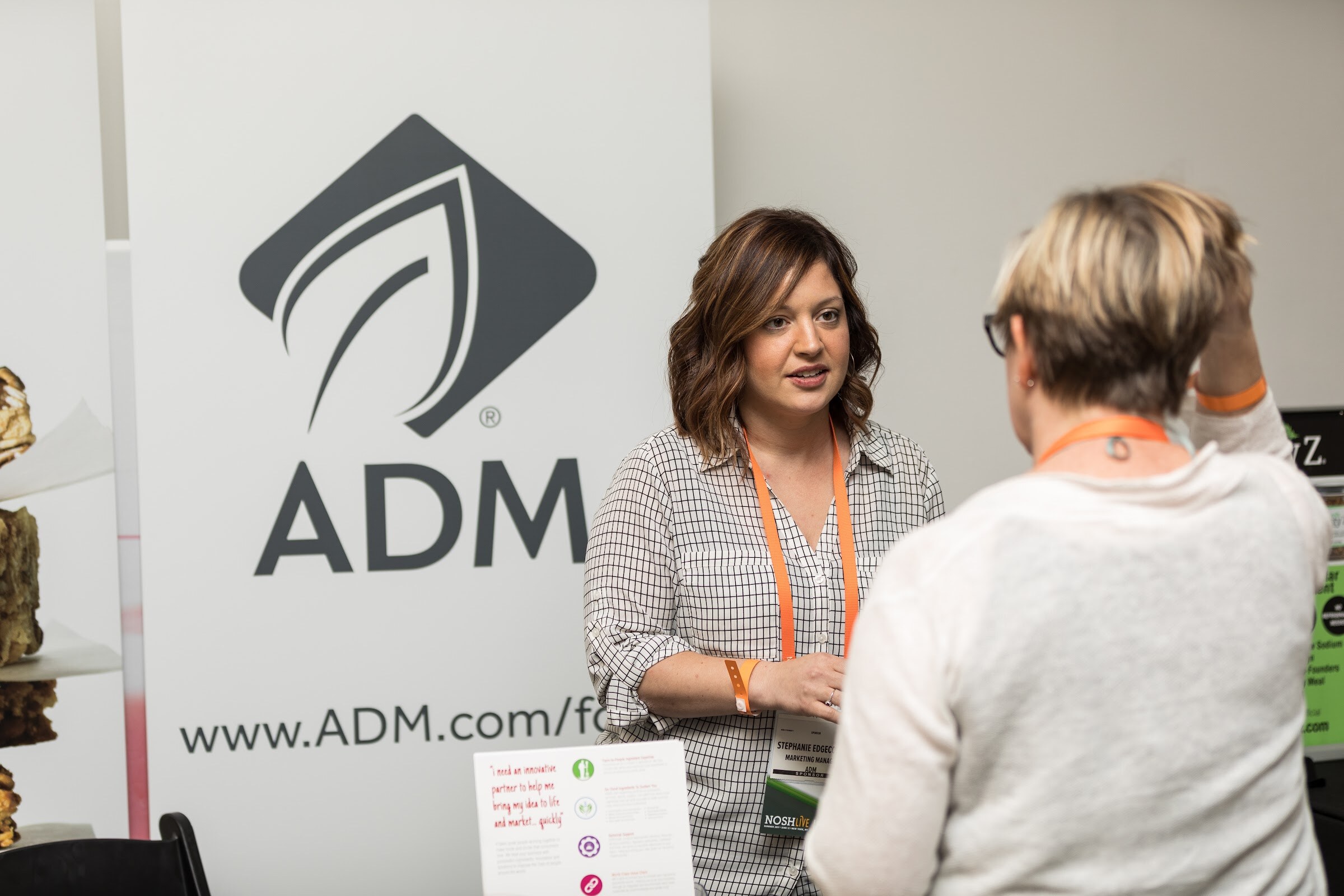 A woman with brown hair in a checked blouse speaks with an attendee in front of a tall ADM banner and a table of snack bars.