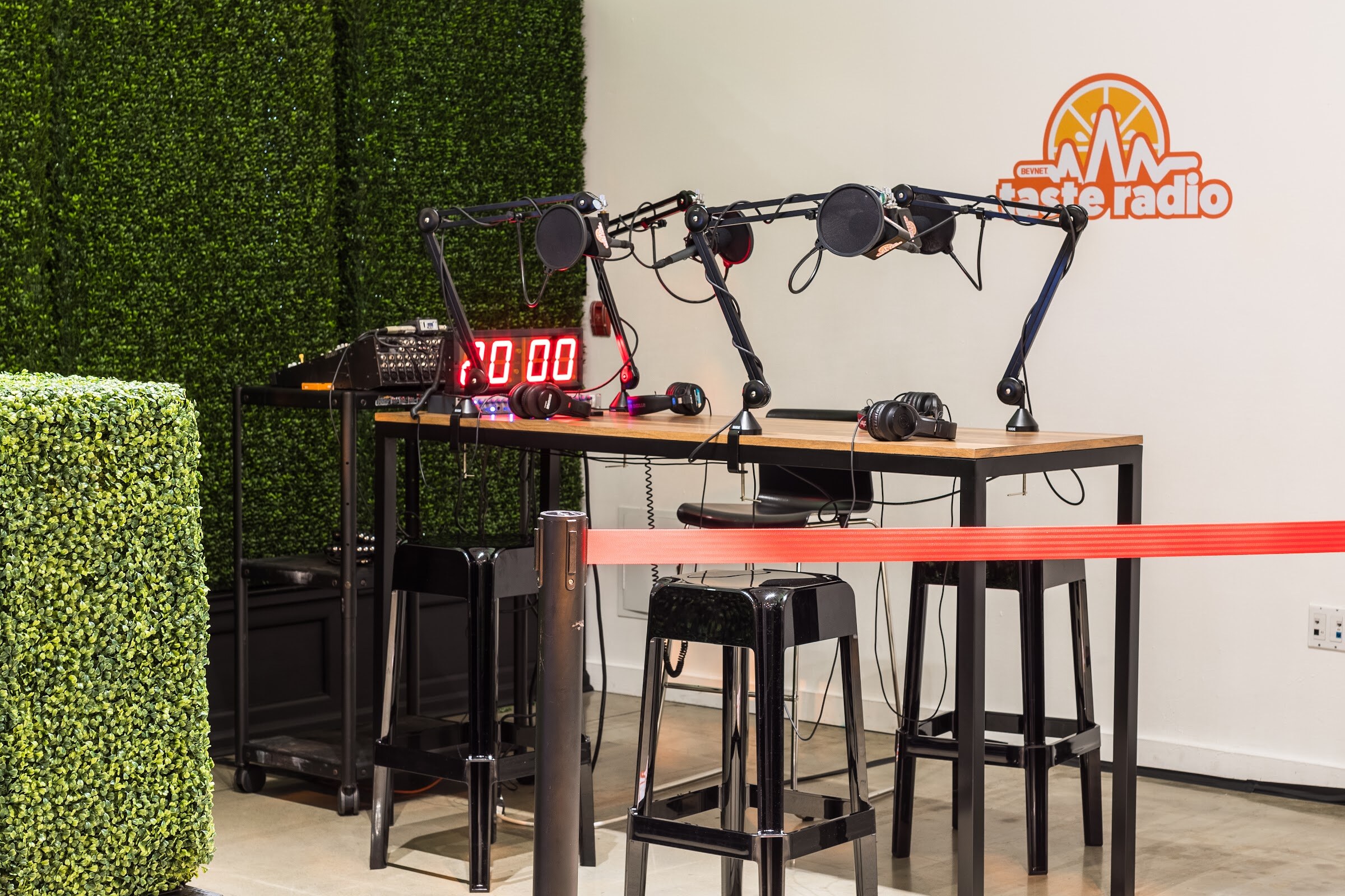 A wooden table with boom-arm microphones, headphones and a red digital timer in front of a hedge wall and a Taste Radio logo on a white wall, closed off with a red ribbon.