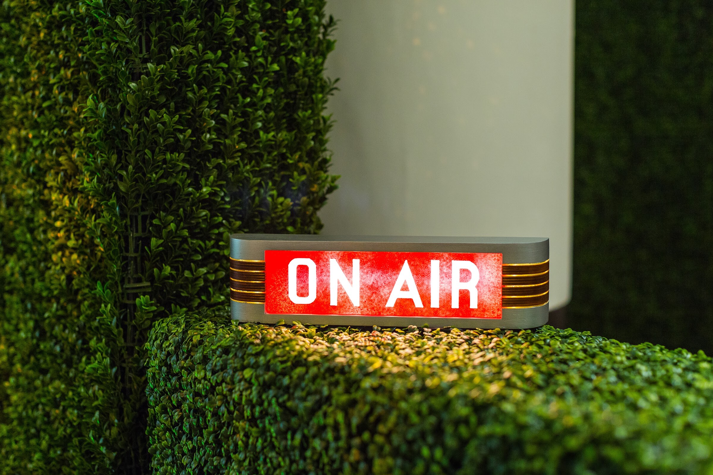 A red illuminated On Air sign sits on top of a green hedge in a studio corner.
