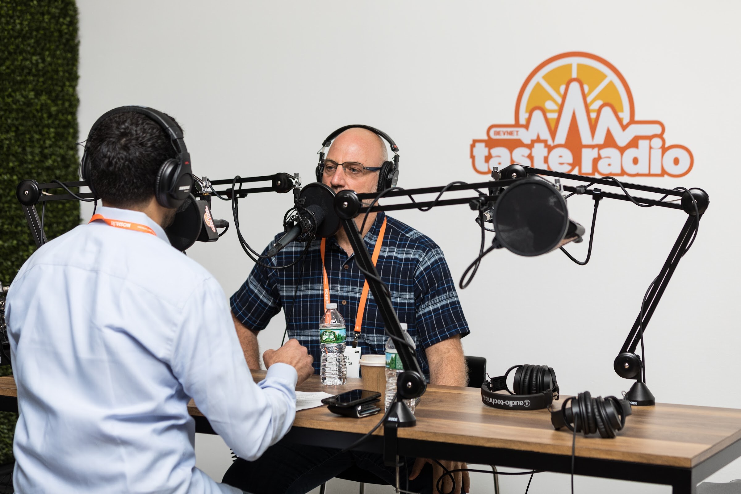 Two men in headphones sit across a wooden table with boom microphones and pop filters in front of an orange Taste Radio logo.