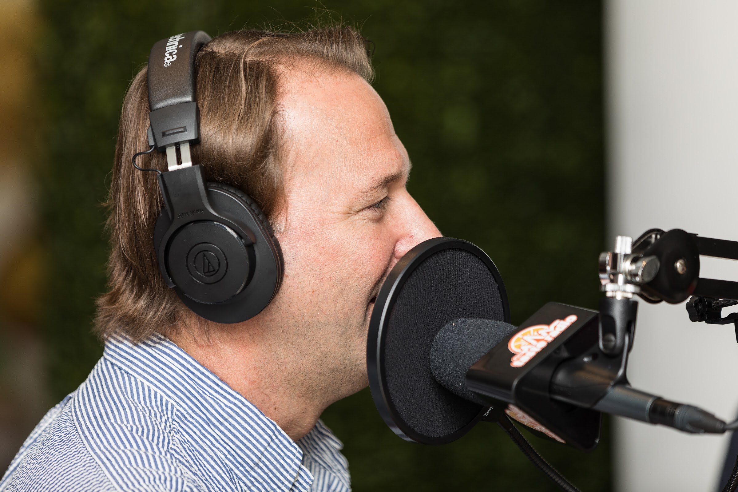 A man in a striped shirt and black headphones speaks into a microphone with a pop filter and a Taste Radio logo.