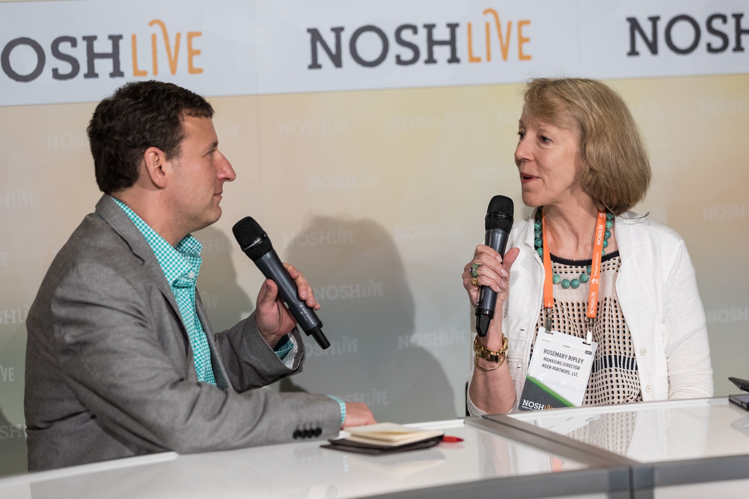 A man in a gray blazer and a woman with an orange lanyard hold microphones and talk across a white desk in front of a Nosh Live banner.