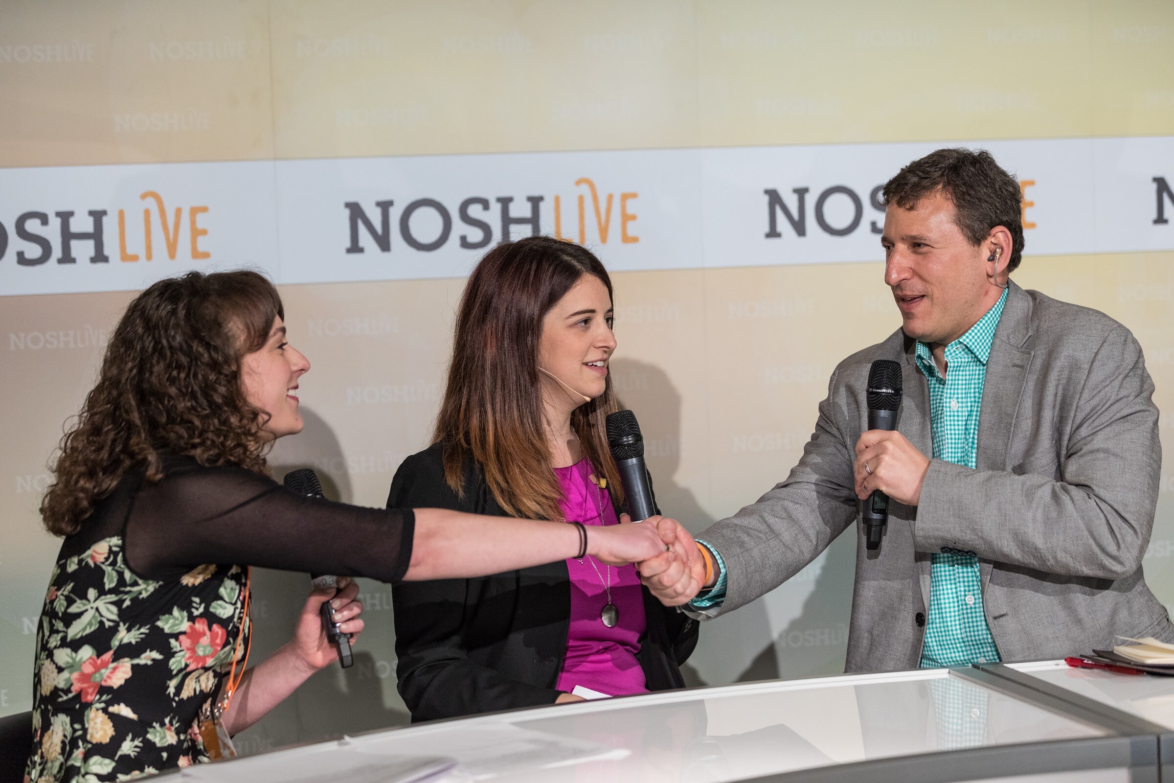 Two women and a man in a gray blazer holding microphones shake hands over a white desk in front of Nosh Live banners.
