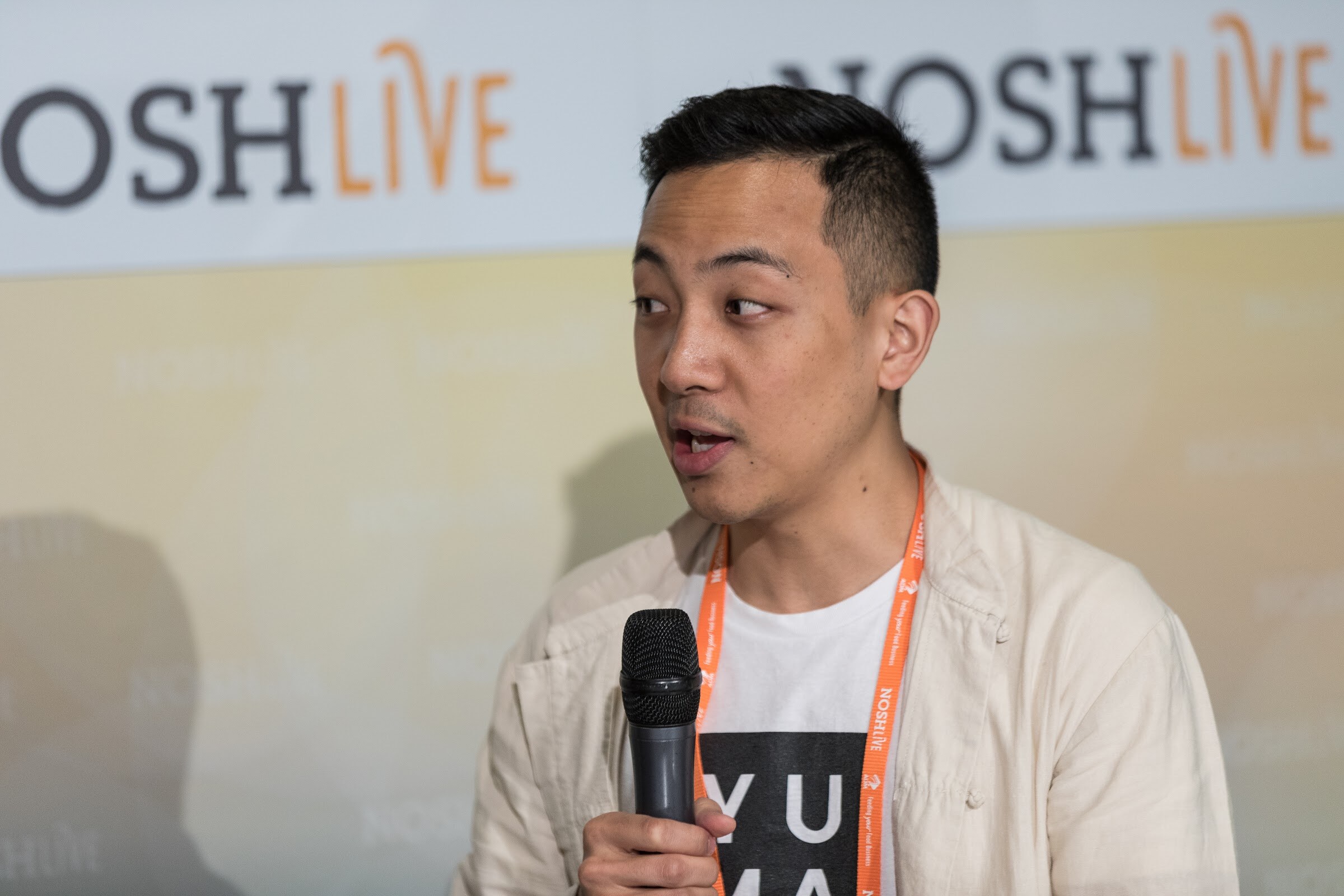A man in a cream jacket and a graphic t-shirt holds a microphone and speaks in front of a Nosh Live banner.