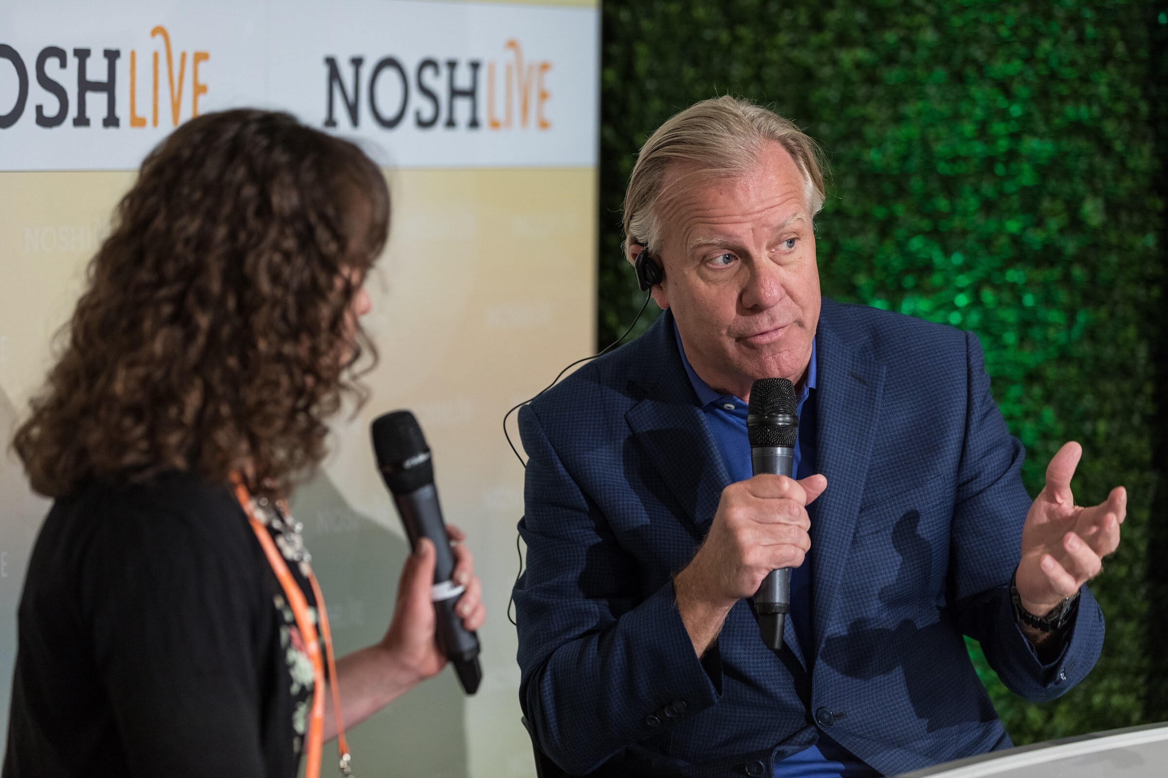 A man in a navy blazer holds a microphone and gestures beside a woman with curly hair, with a hedge wall behind him.