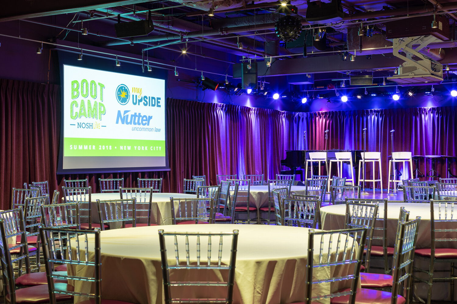 Boot Camp room set with a projected NOSH Boot Camp Summer 2018 slide, round tables and a lit stage with four stools.
