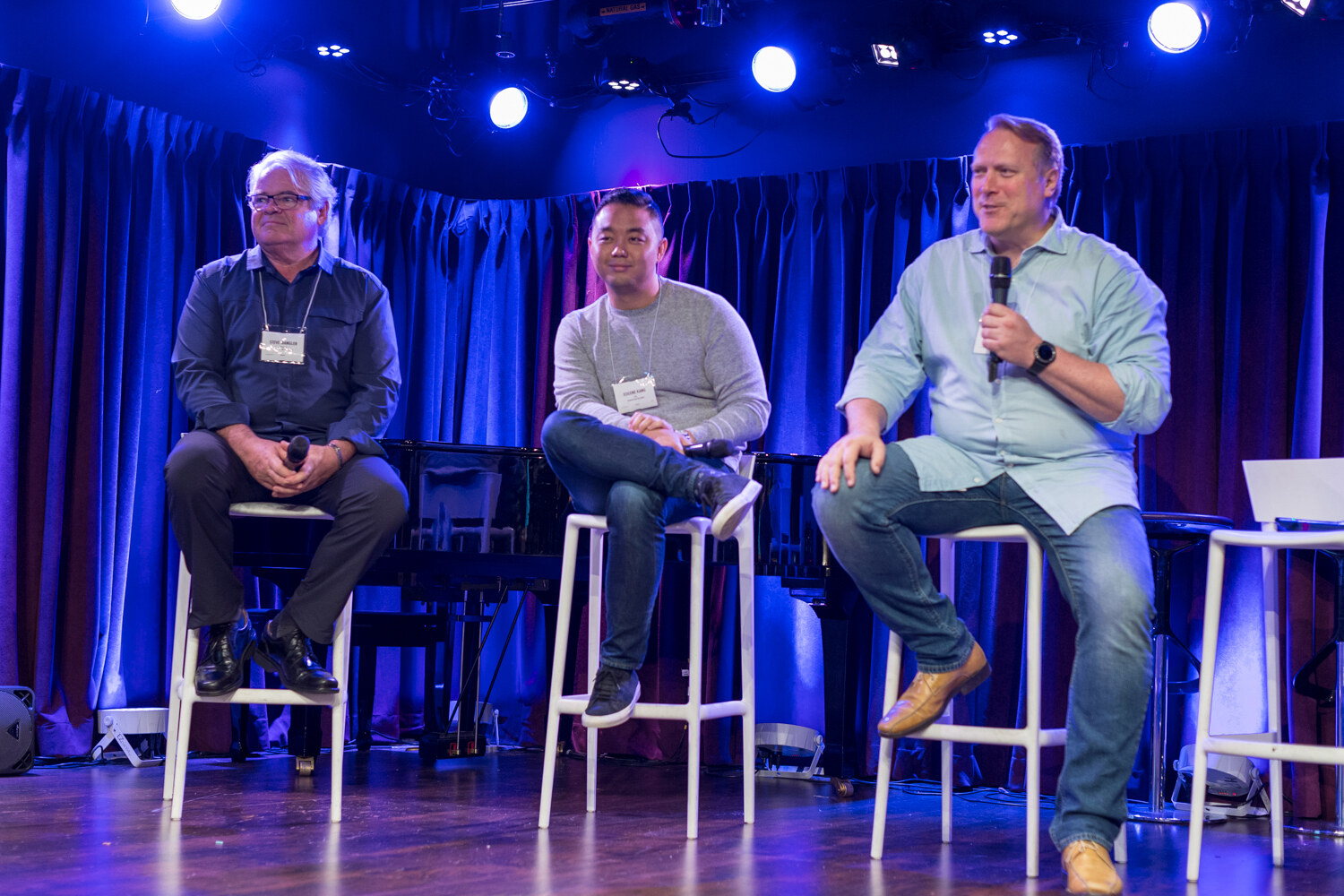 Three panelists seated on stools on a purple-lit stage during the Competing session.