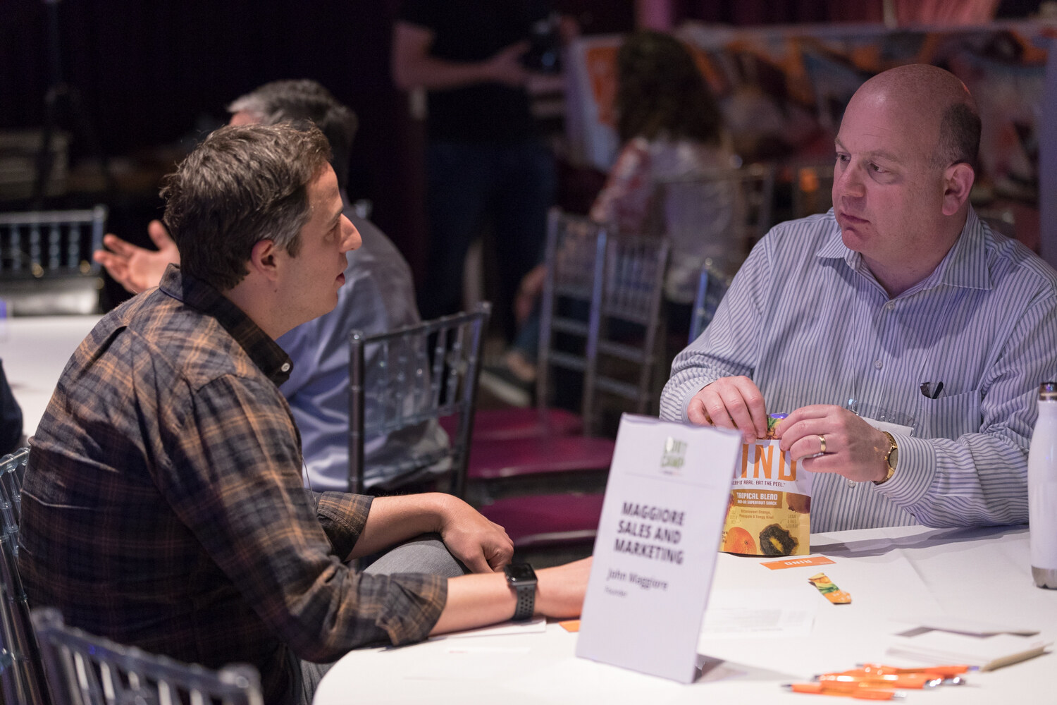 Two people talk at a Boot Camp expert table with a Maggiore Sales and Marketing card and a bag of snacks.