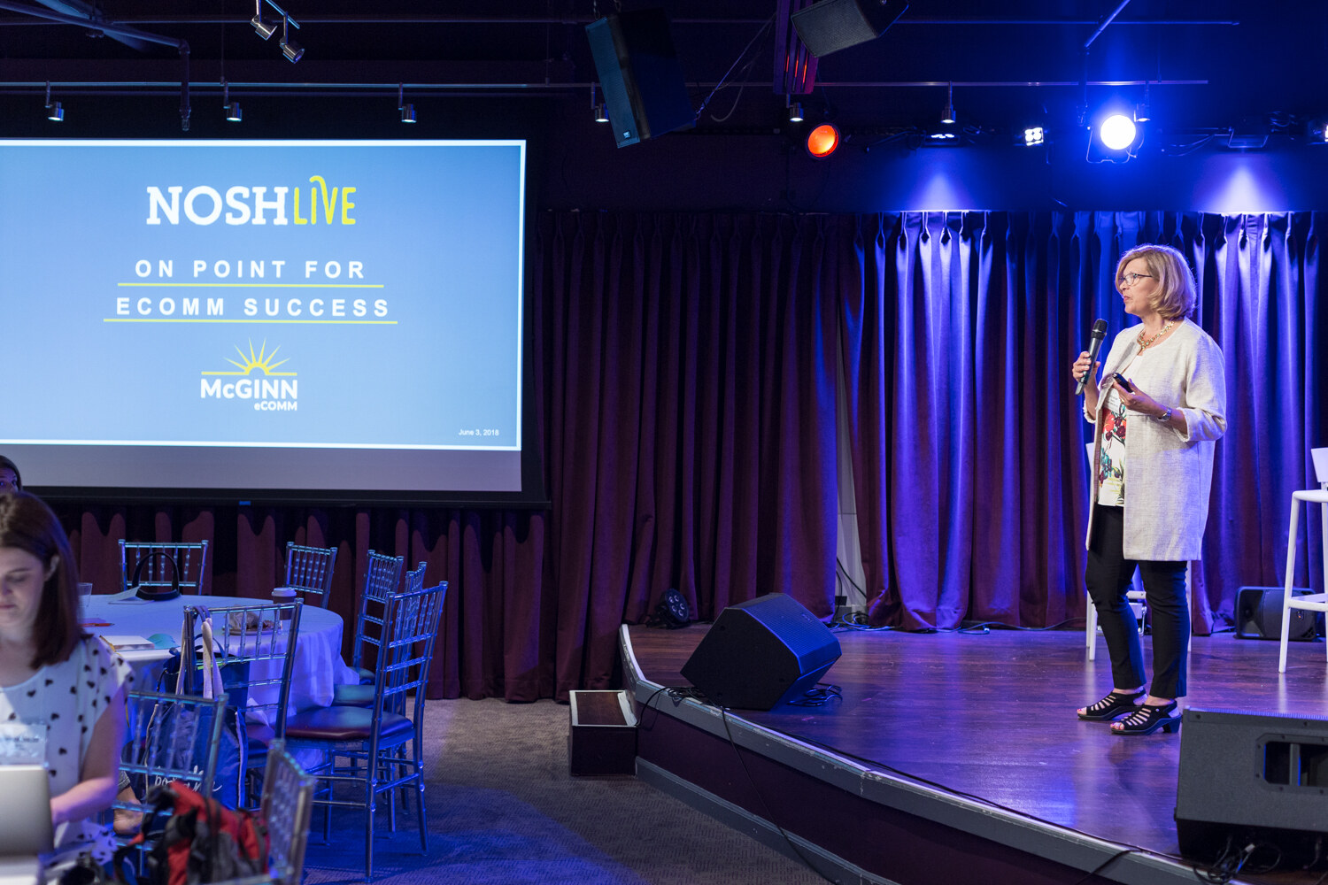 Presenter on stage beside a projected NOSH Live On Point for Ecomm Success slide with a McGinn logo.