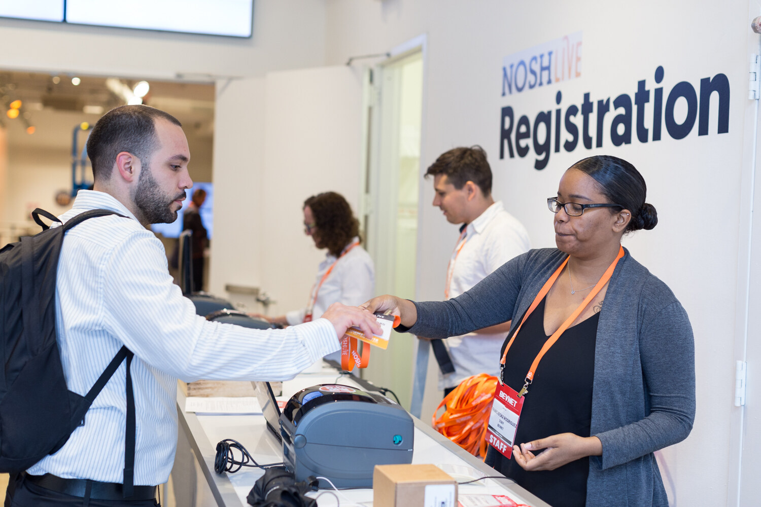 A registration staffer hands a lanyard and badge to an attendee at the Nosh Live check-in desk.