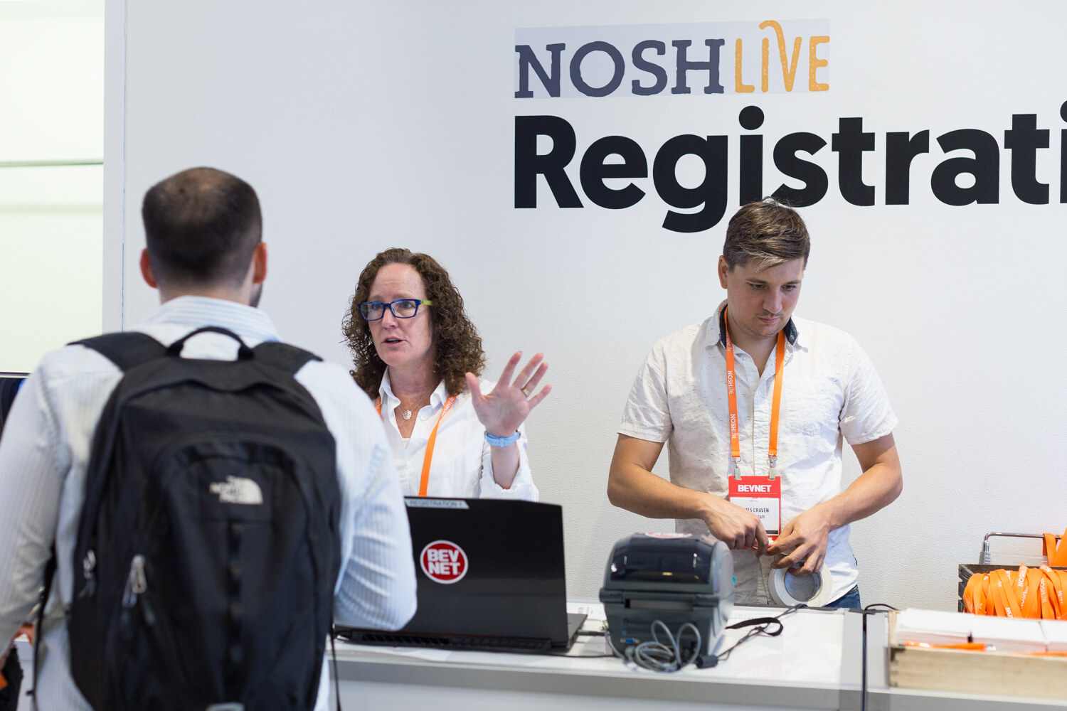 Registration staff and an arriving attendee at the Nosh Live registration desk with a laptop and badge printer.
