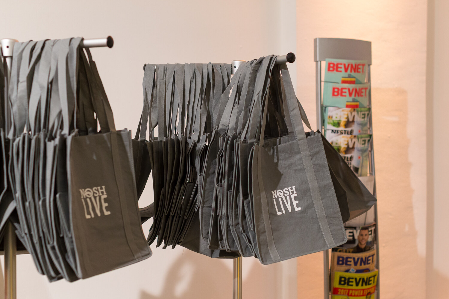 Gray Nosh Live tote bags hang on a rack beside a rack of BevNET magazines.