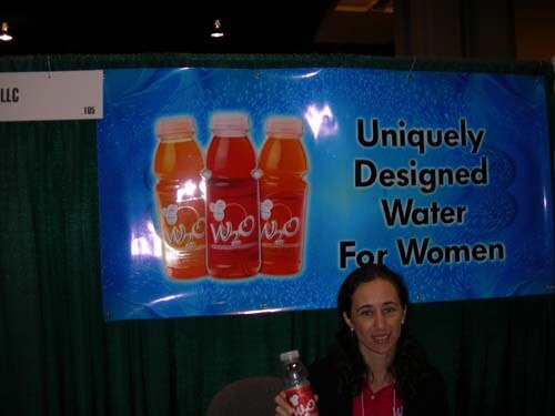 A woman sits behind a W2O booth table holding a bottle of W2O, under a blue banner reading Uniquely Designed Water For Women with three bottles pictured.