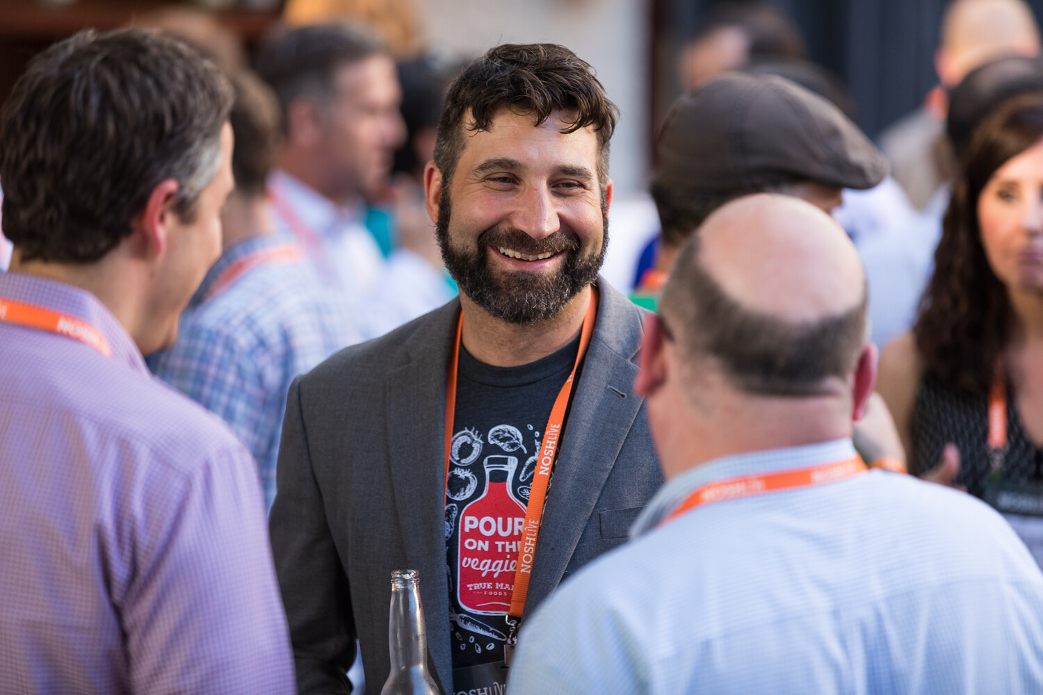 A man in a gray blazer and graphic t-shirt smiles in the crowd at the Nosh Live afterparty.