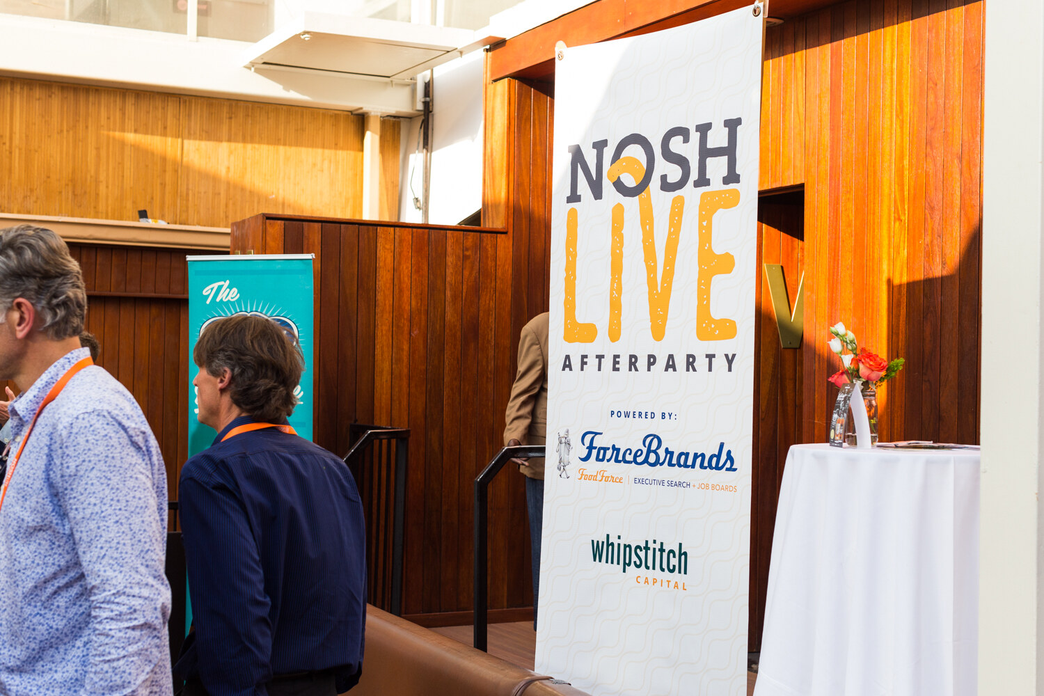 A tall NOSH LIVE Afterparty banner powered by ForceBrands stands beside a wooden wall as guests talk.