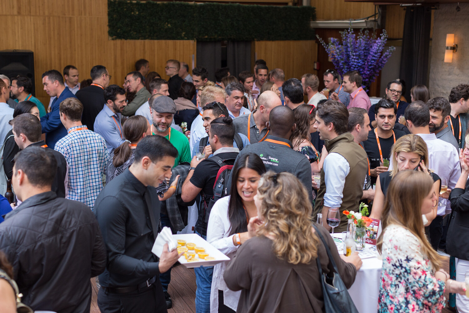 A crowd of guests fills the wooden-paneled terrace at the Nosh Live afterparty while a server passes a tray.
