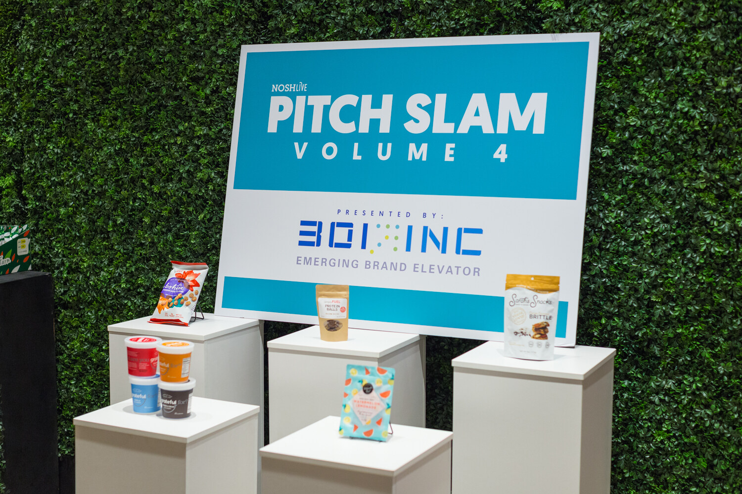 A Pitch Slam Volume 4 sign presented by 301 Inc displayed on an ivy wall above pedestals of products.