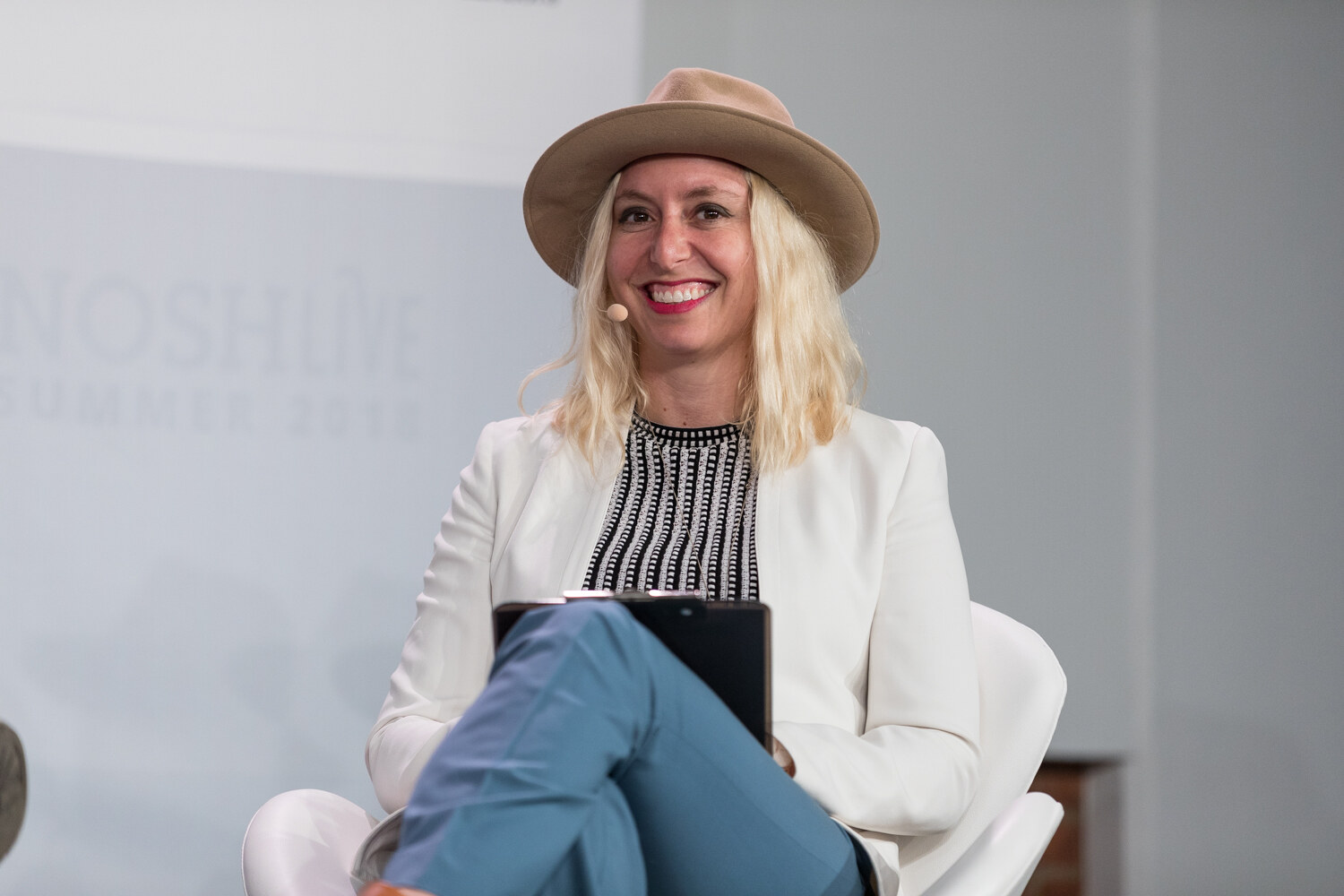 A Pitch Slam judge in a tan hat and white blazer smiles while seated with a tablet on her lap.