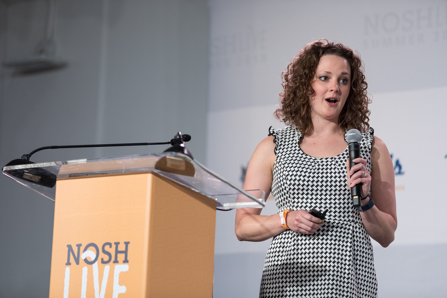 A host in a houndstooth dress holds a microphone beside the NOSH LIVE lectern while introducing Pitch Slam.