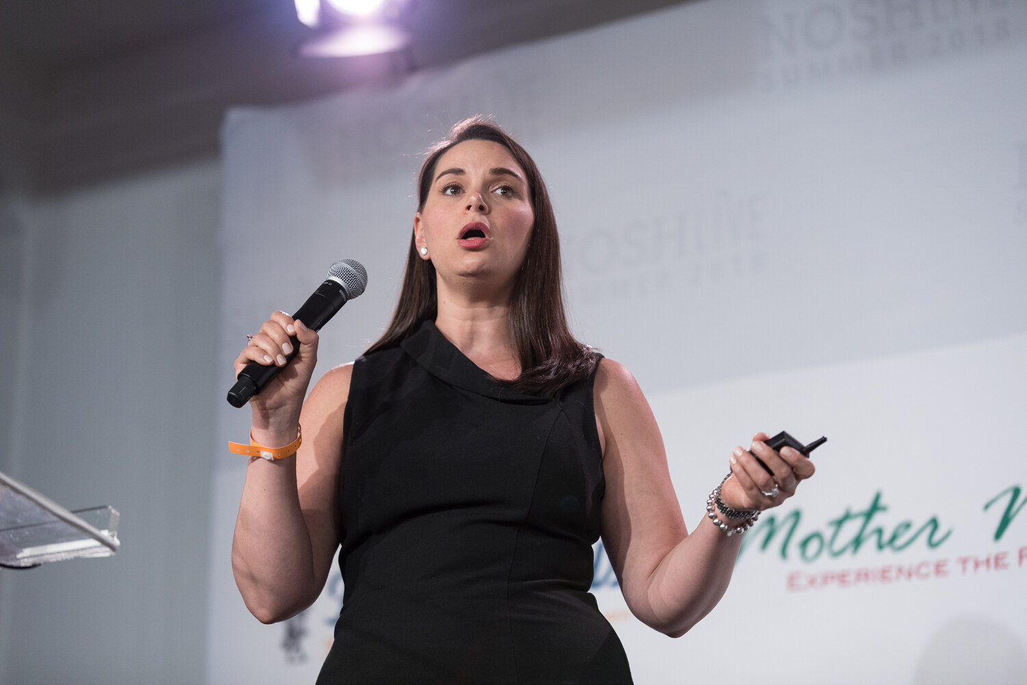 A Pitch Slam contestant in a black dress gestures with a microphone and clicker on stage.