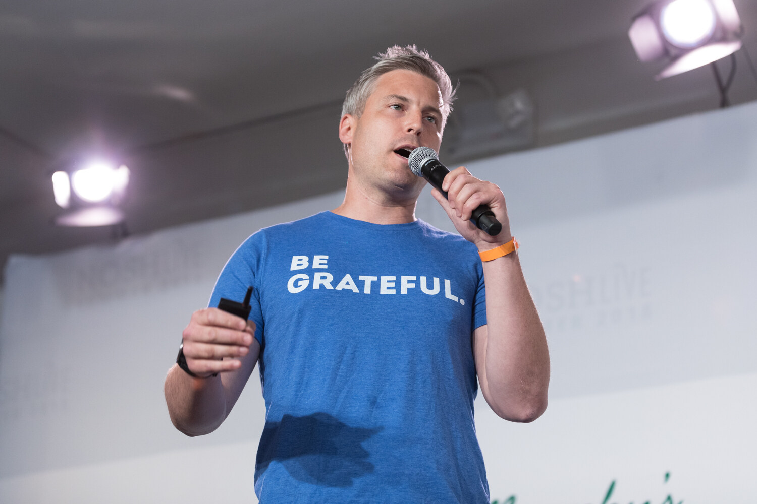 A Pitch Slam contestant in a blue Be Grateful t-shirt speaks into a microphone on stage.