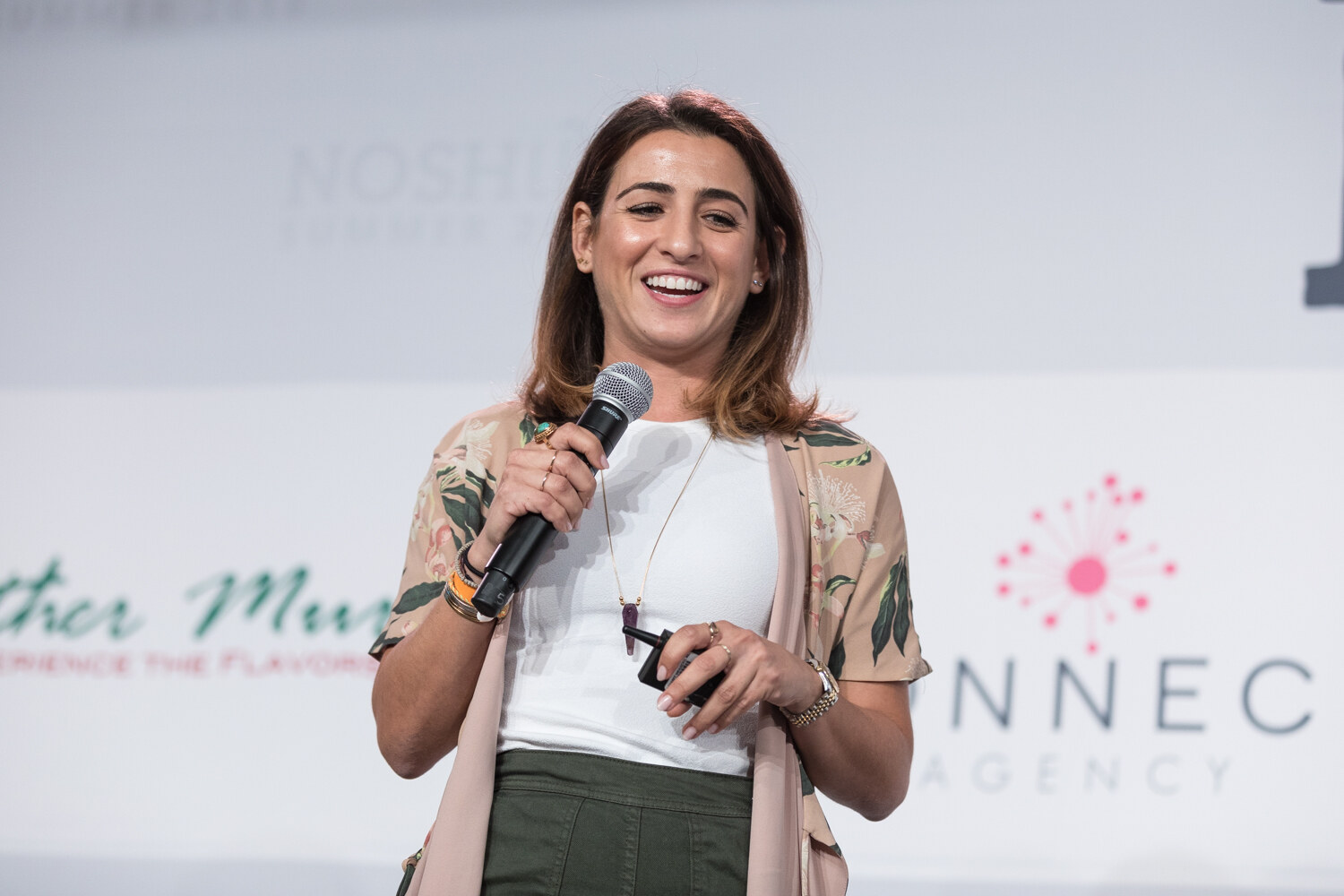 A Pitch Slam contestant in a floral duster jacket laughs while holding a microphone in front of a Konnect Agency banner.