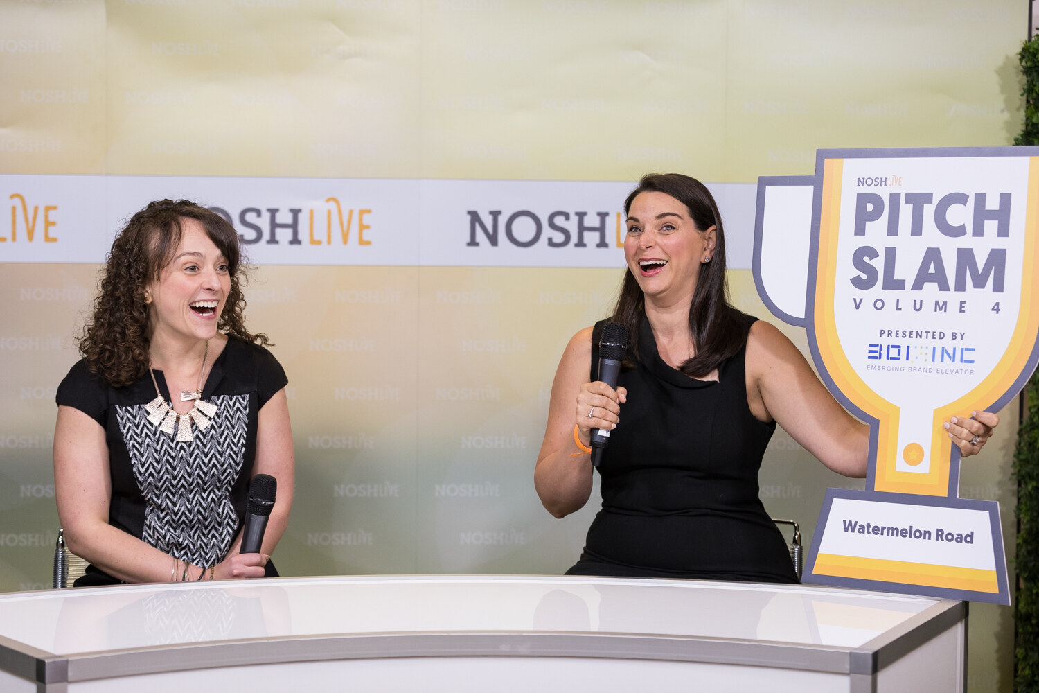 Two women laugh at the Nosh Live livestream desk beside a Pitch Slam Volume 4 trophy sign reading Watermelon Road.