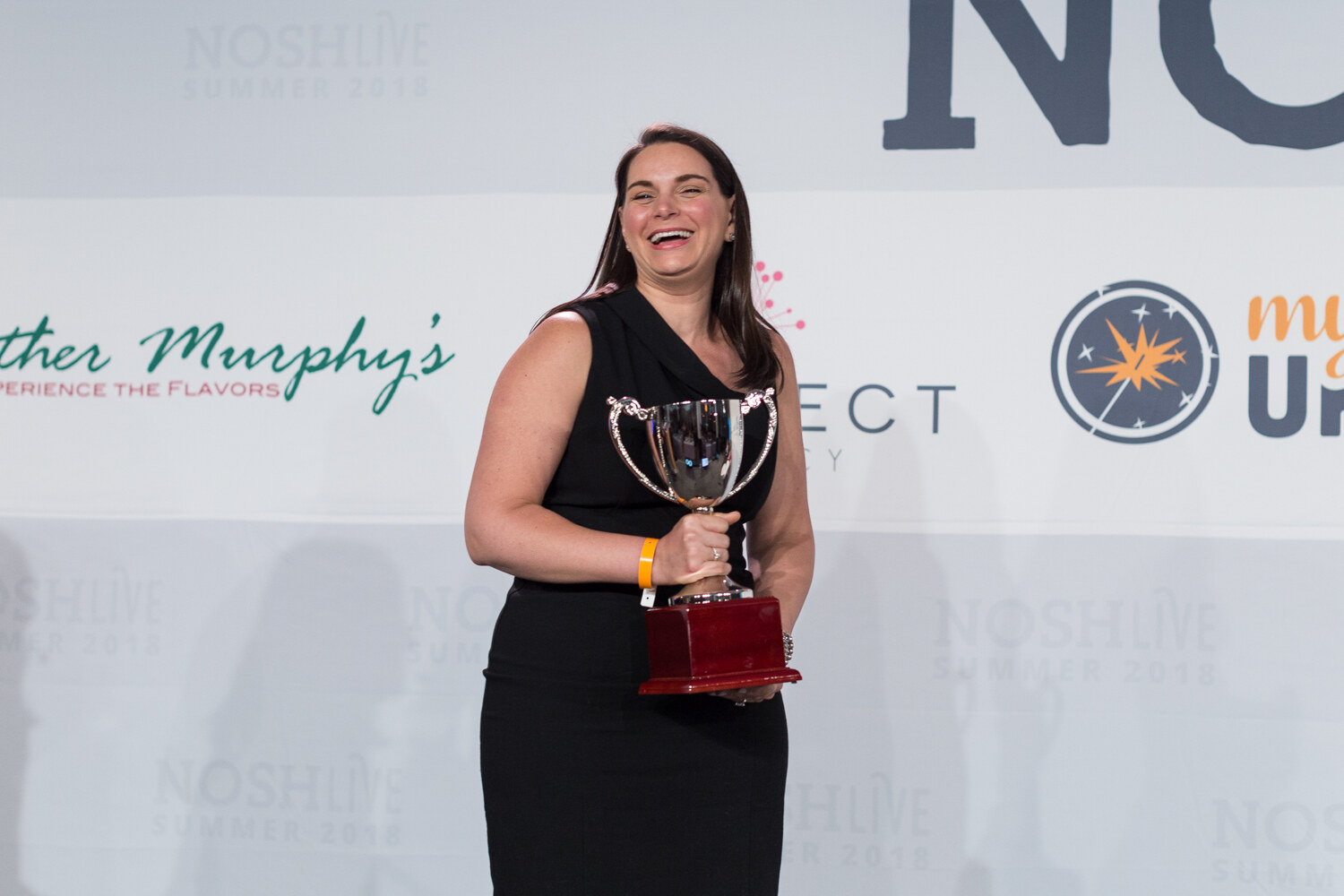 A woman in a black dress laughs while holding a silver trophy cup on the Nosh Live stage.
