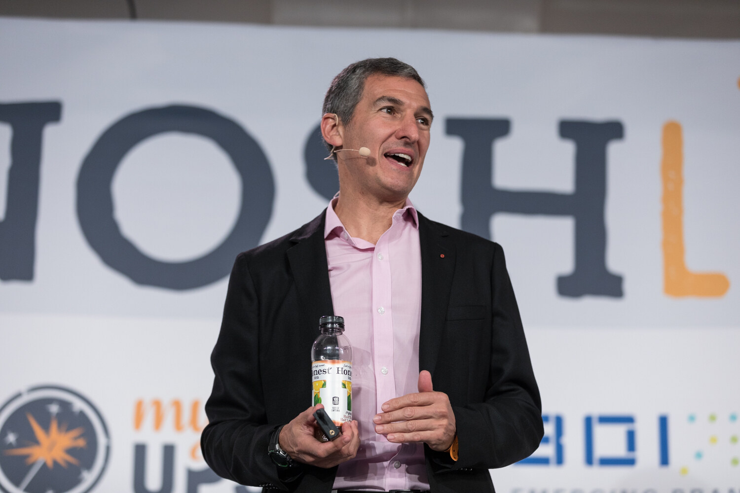 A speaker holding a bottle smiles on stage in front of the NOSH Live backdrop during the Leadership Notes from the Chairman and TeaEO session.