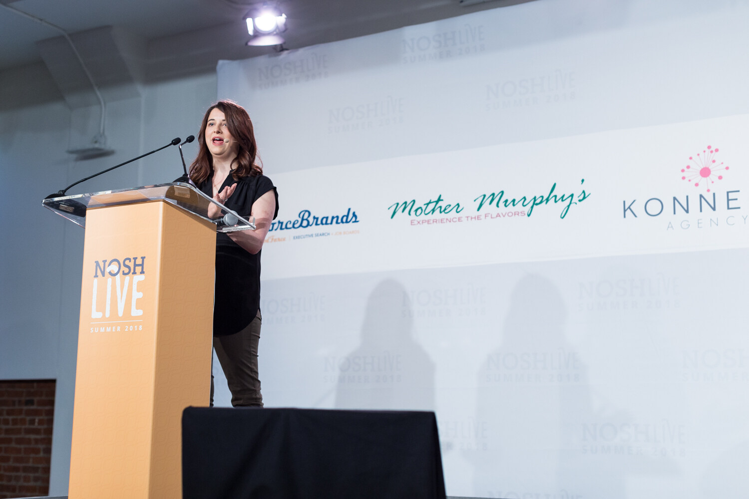 Wide view of a speaker at the NOSH Live lectern in front of a sponsor backdrop during the welcome remarks.