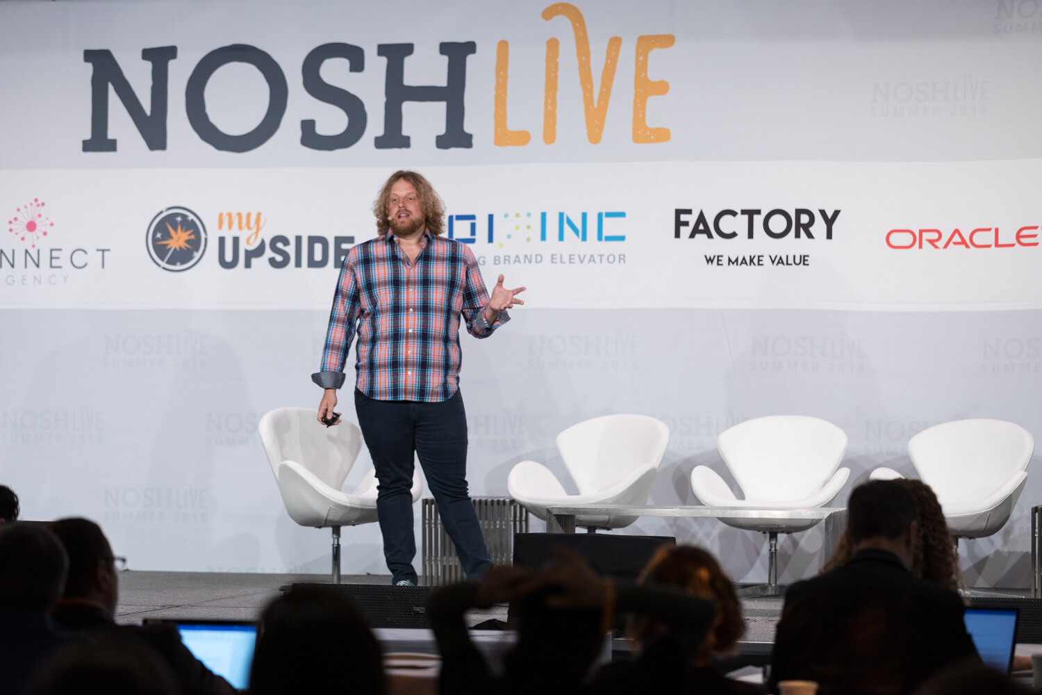 A presenter in a plaid shirt on the Nosh Live stage in front of a seated audience during the Thriving Brands session.