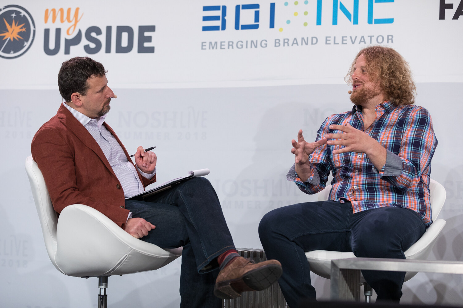 A moderator in a rust blazer and a guest in a plaid shirt talk in white chairs during the Thriving Brands session.
